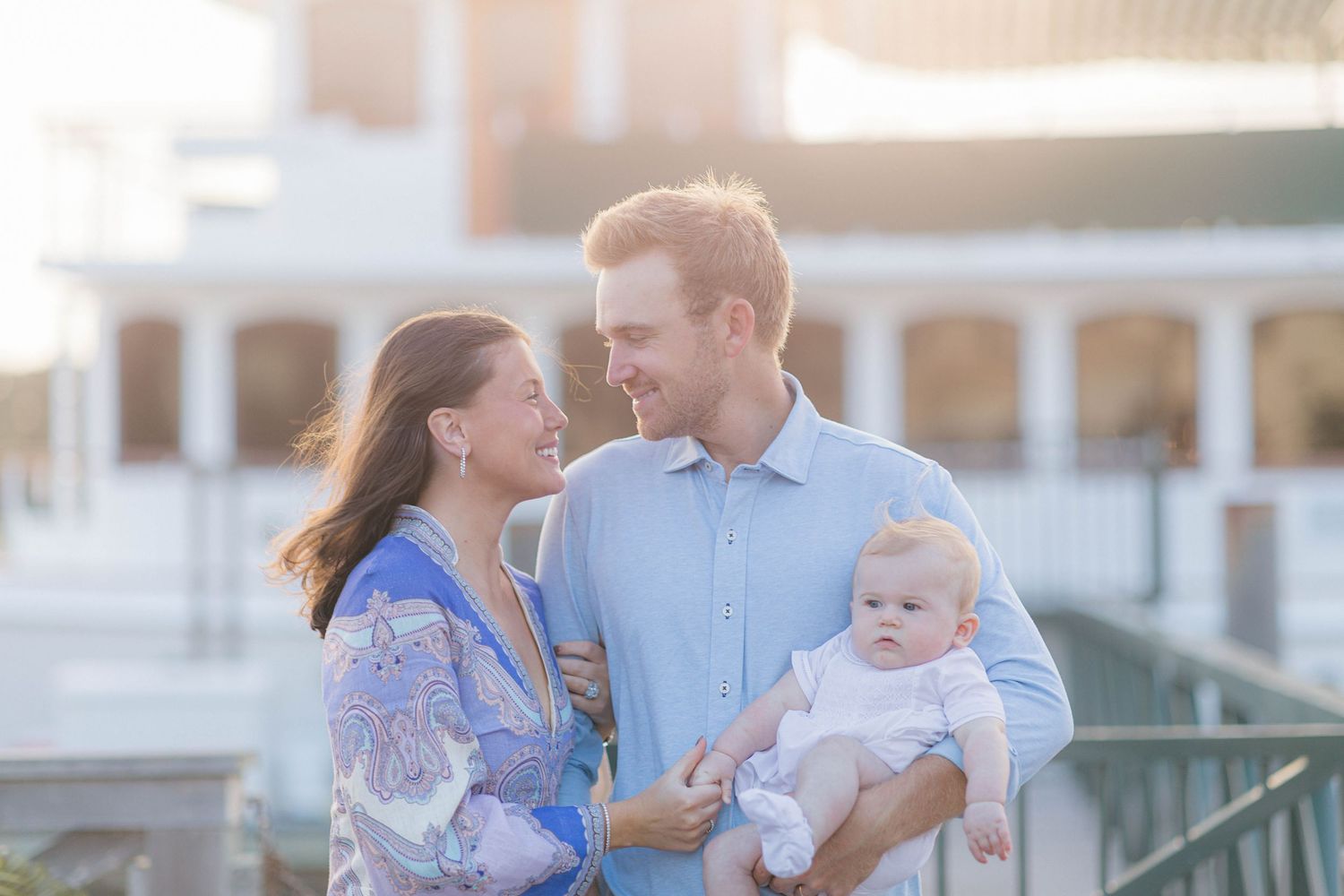 A young family smiles together outdoors, with a man holding a baby in white and a woman in a blue floral dress.