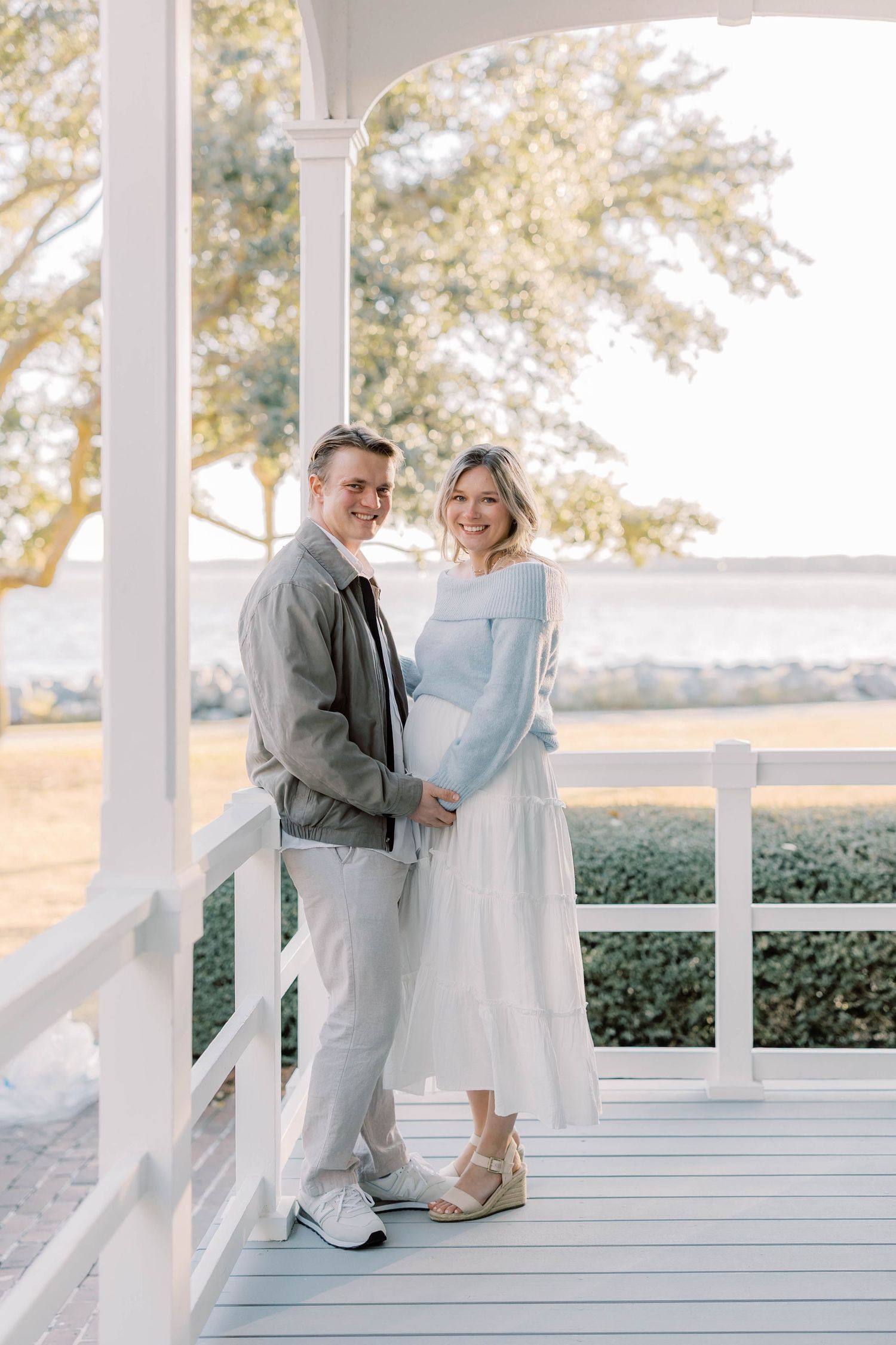 Couple poses on white porch, woman in blue sweater and white dress cradles baby bump beside man in gray jacket.