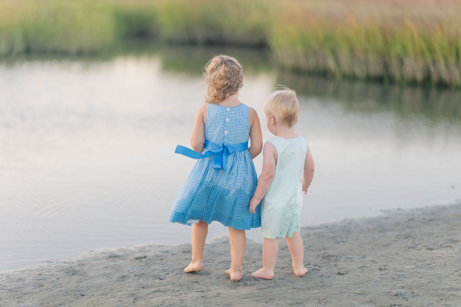 Two young girls in blue and mint dresses stand together at the water's edge on a sandy beach.