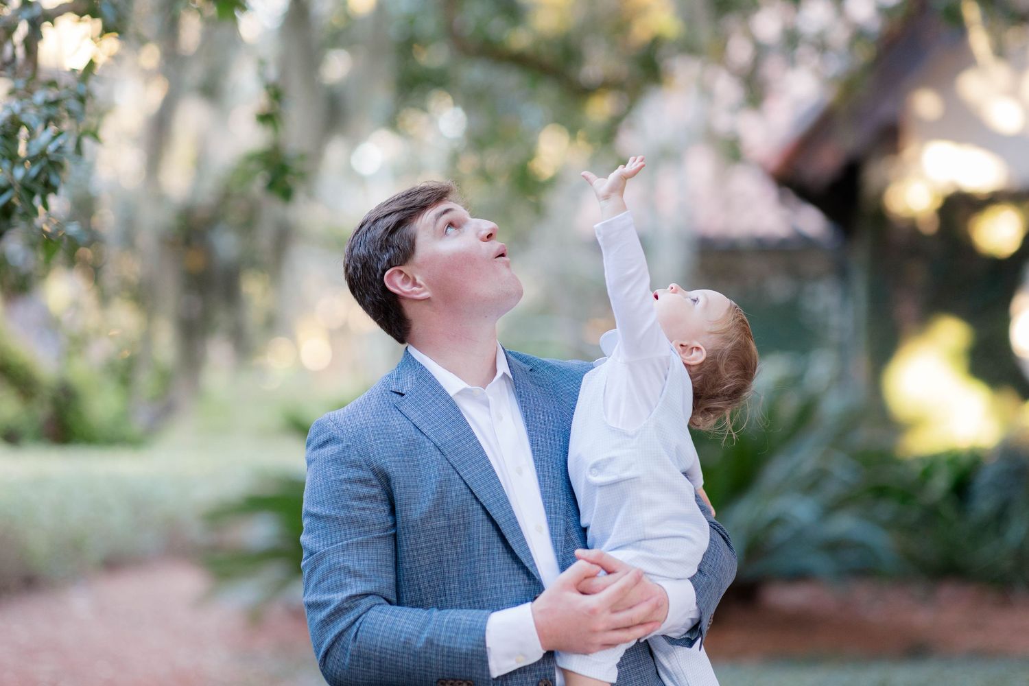 A father in a gray suit holds his baby outdoors, both looking up at the sky among lush trees.