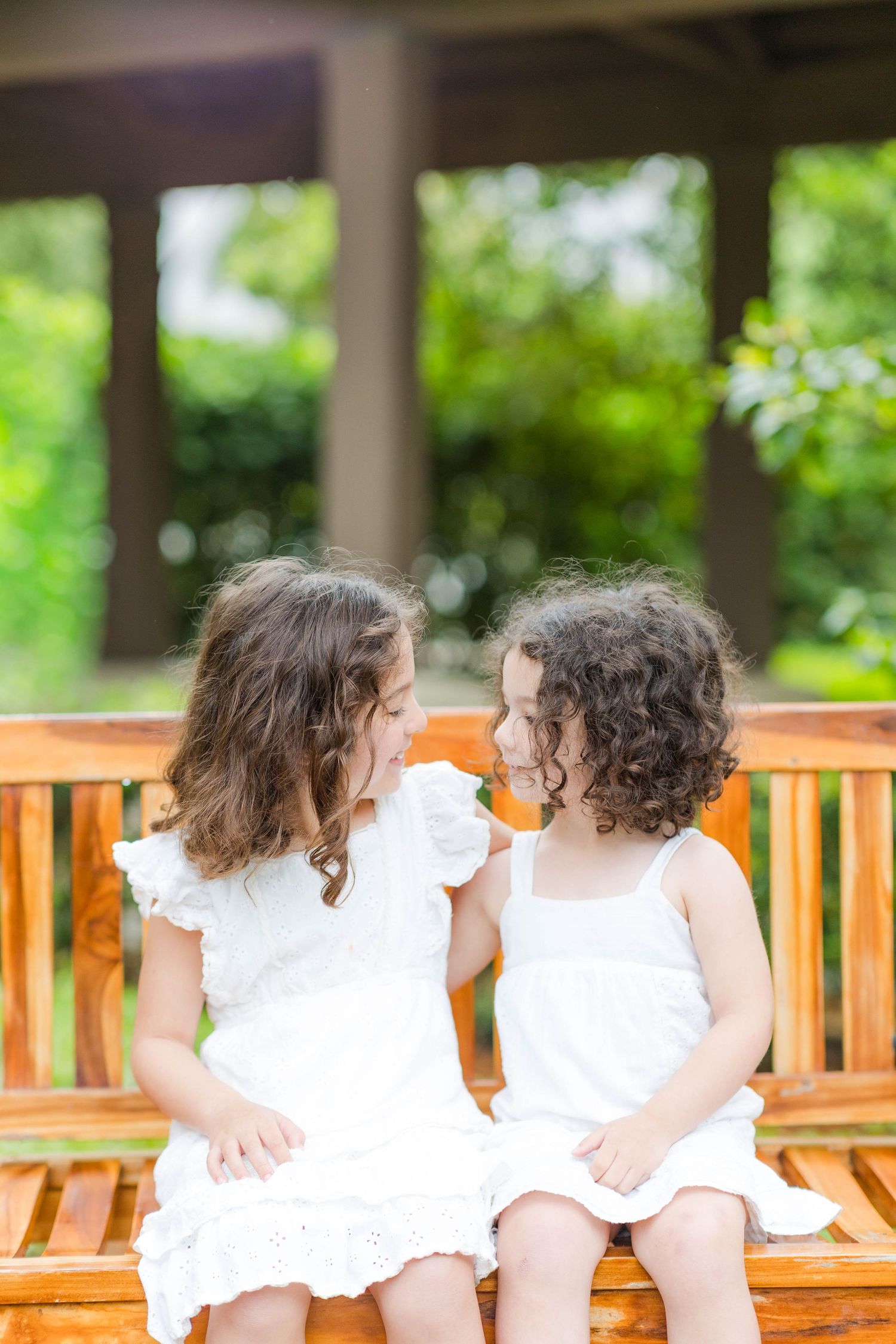 Two young girls in white dresses sitting on a wooden bench, smiling at each other outdoors.