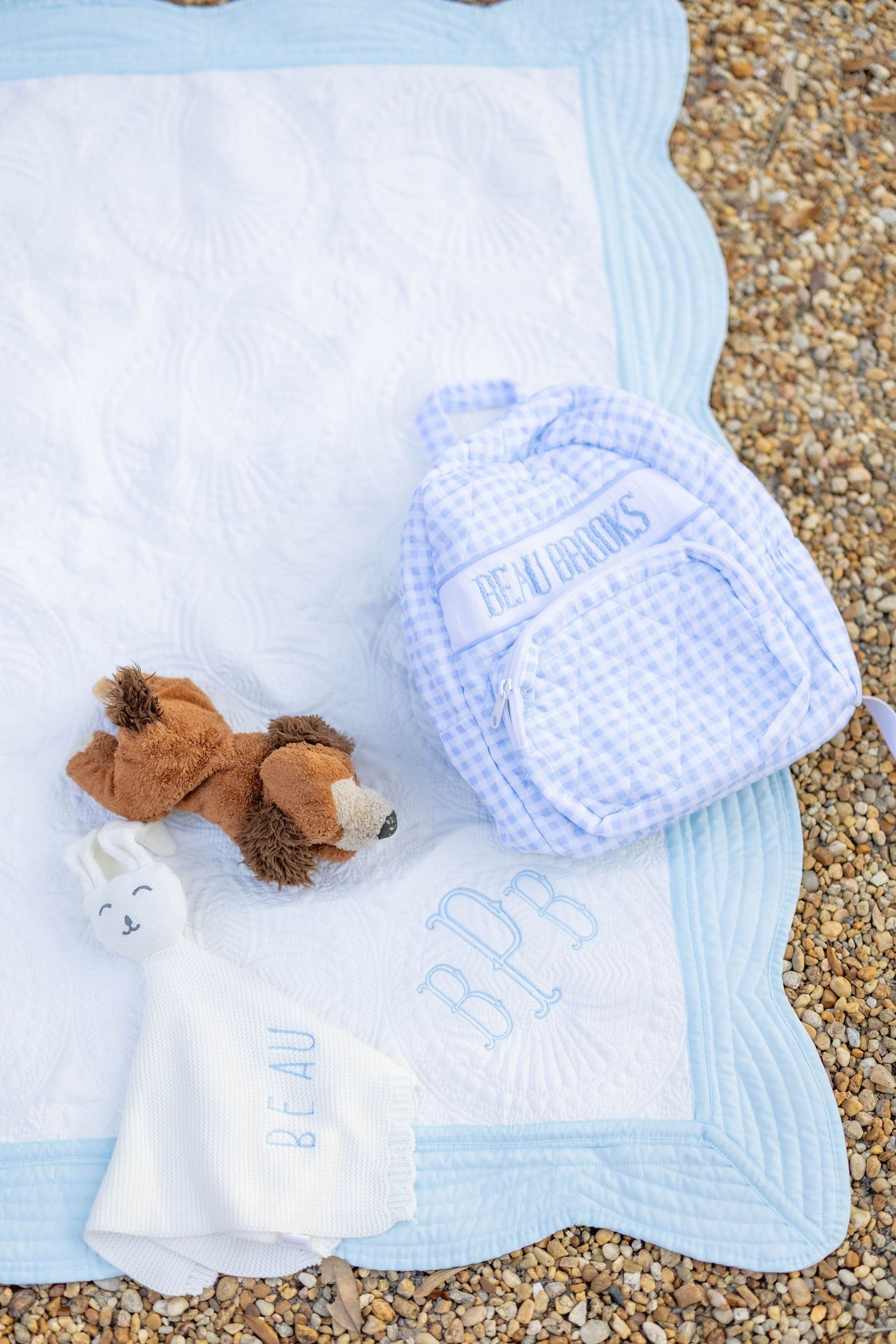Baby quilt with monogram, gingham backpack, stuffed bear, and personalized lovey blanket on sandy beach.