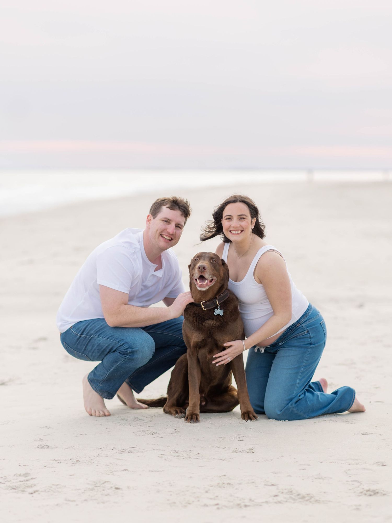 A couple in white and denim sits on a sandy beach with their chocolate Labrador dog during a casual photoshoot.
