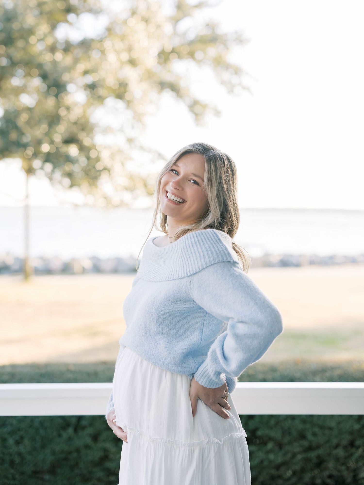 Smiling pregnant woman in light blue sweater and white skirt poses outdoors near white fence on sunny day.