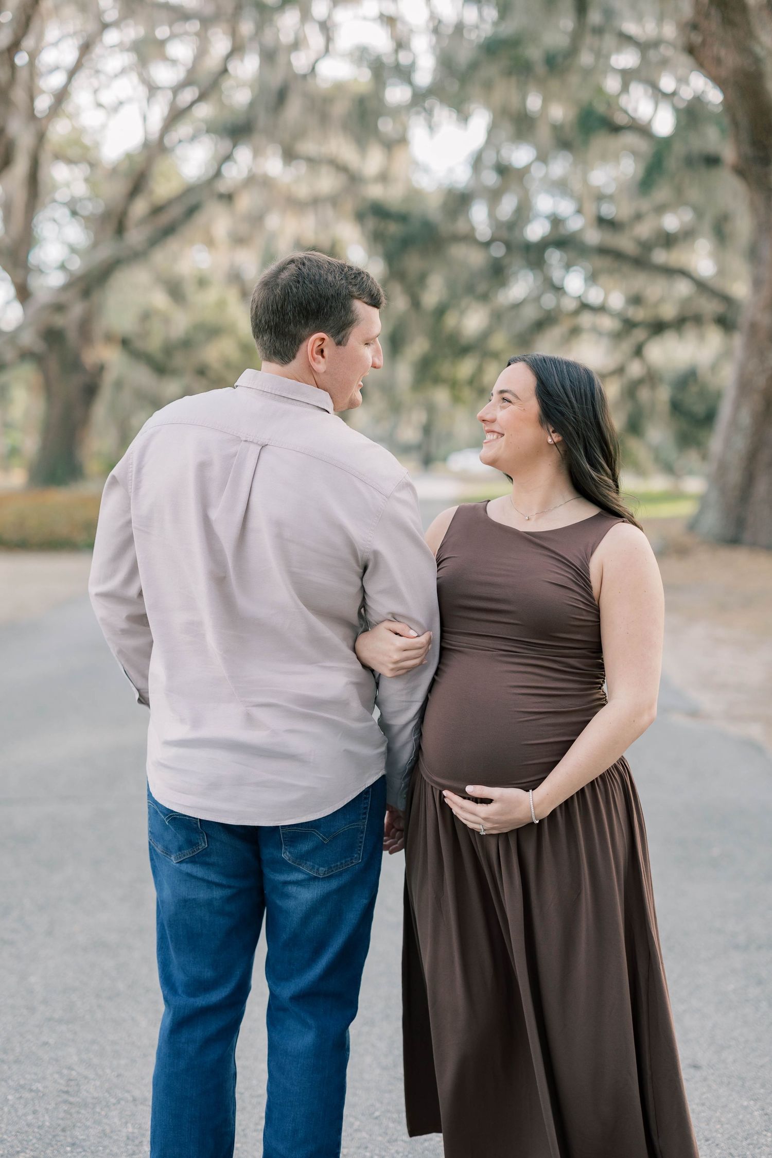 Pregnant couple smiling at each other outdoors, woman in brown maternity dress cradling baby bump, man in gray shirt and jeans.