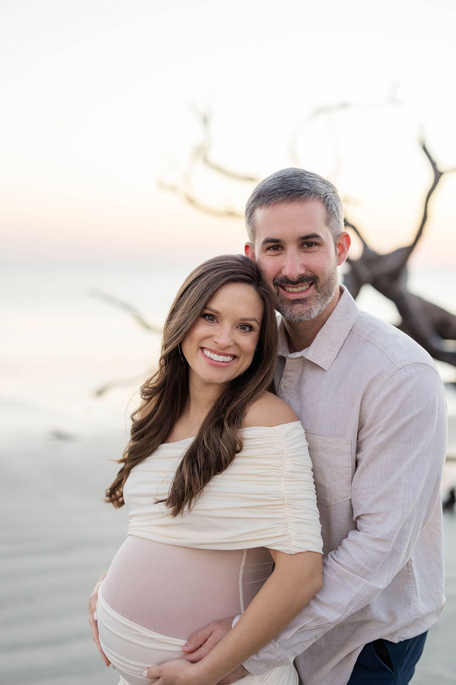 Smiling couple poses on beach at sunset, pregnant woman in white off-shoulder dress, man in light pink shirt.