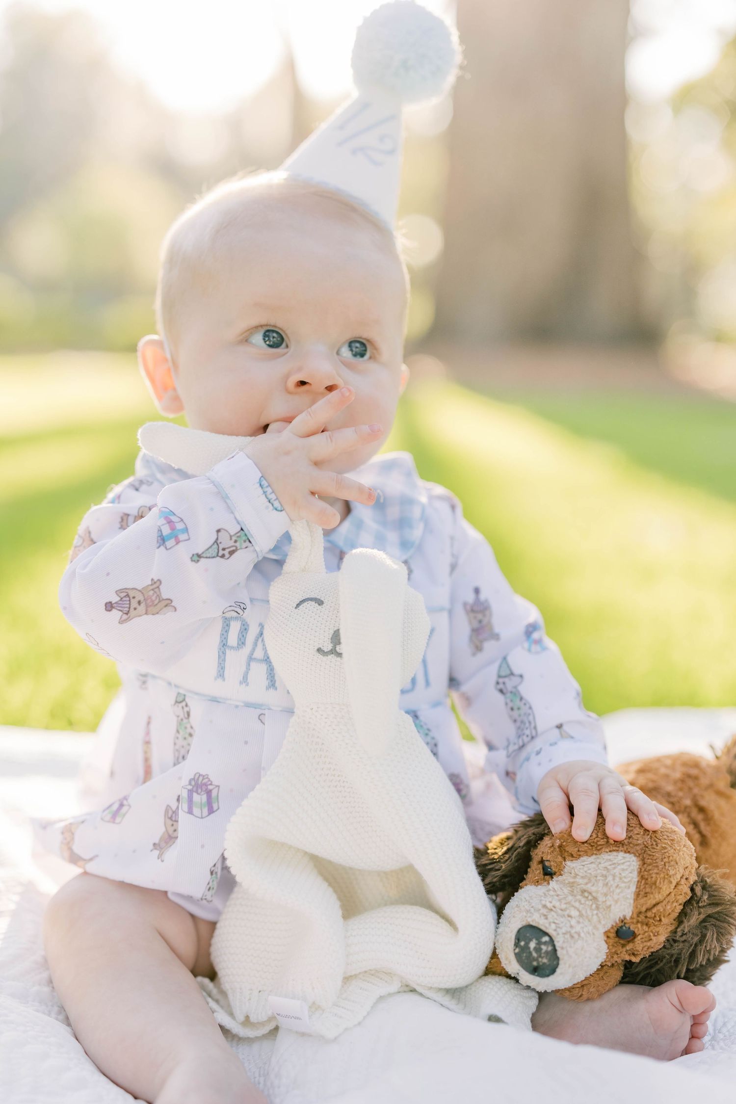 Baby wearing a party hat and printed jacket chews a white stuffed bunny outdoors on a sunny day.