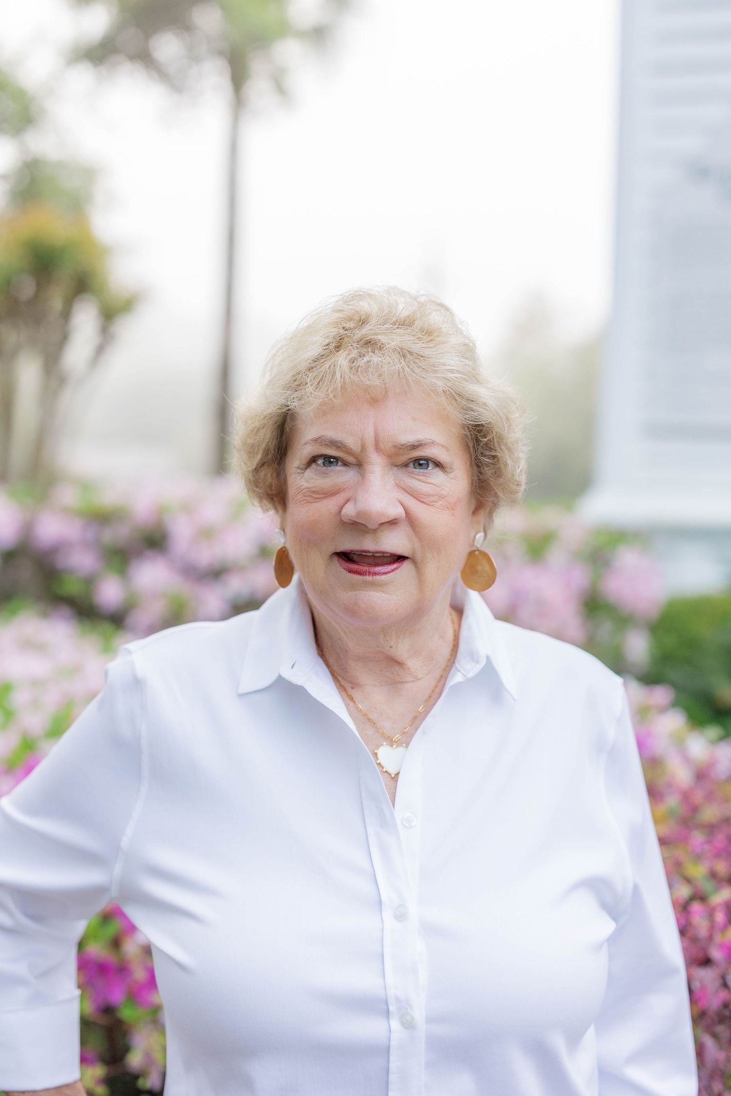 Senior woman in white blouse with orange earrings smiling outdoors near pink flowering bushes.