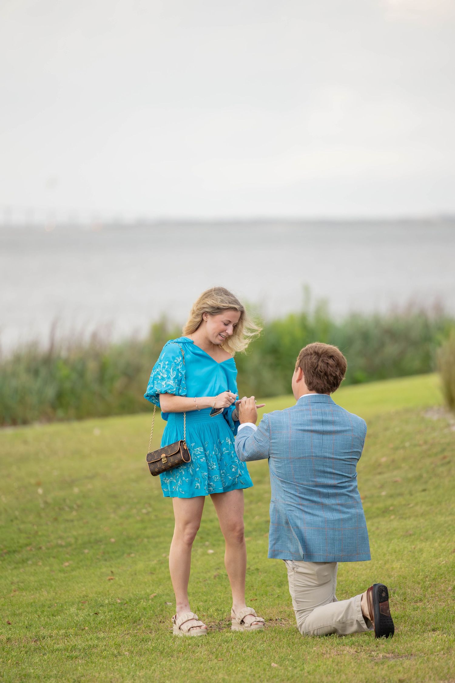 Man kneeling on one knee proposing to a woman in a blue dress outdoors near a waterfront on green grass.