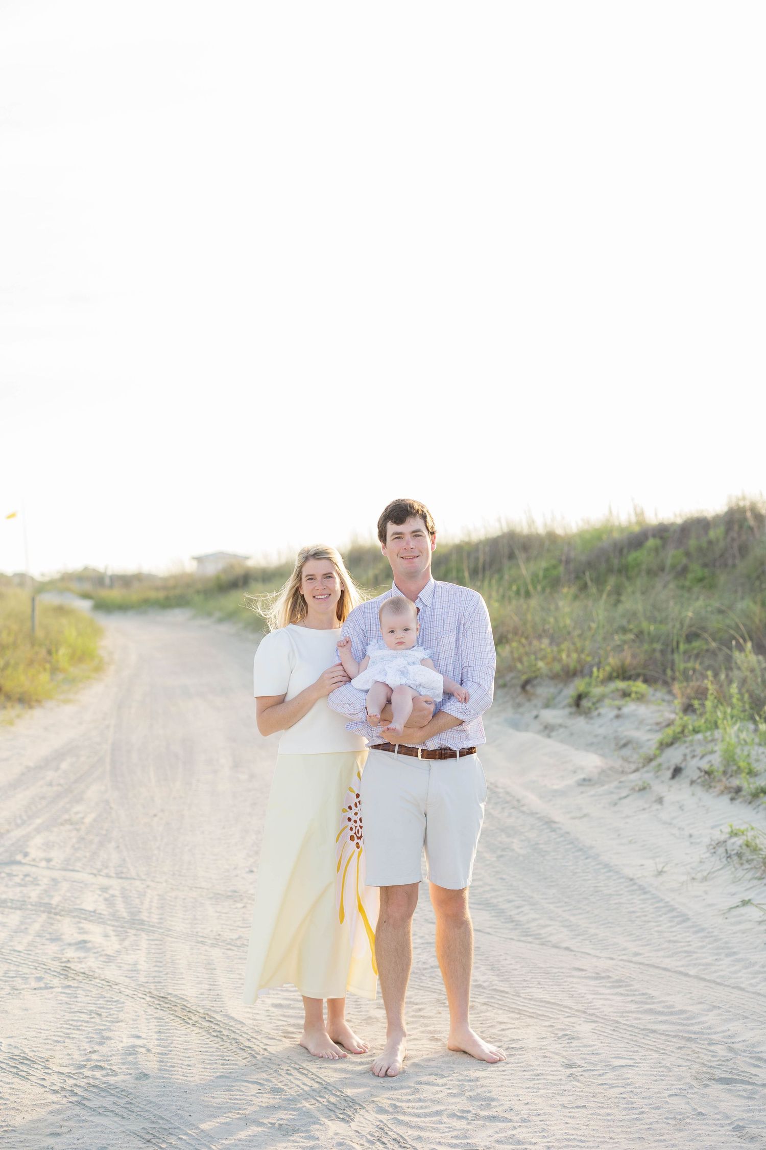 Young family with baby poses on sandy beach path at golden hour, wearing light pastel outfits.