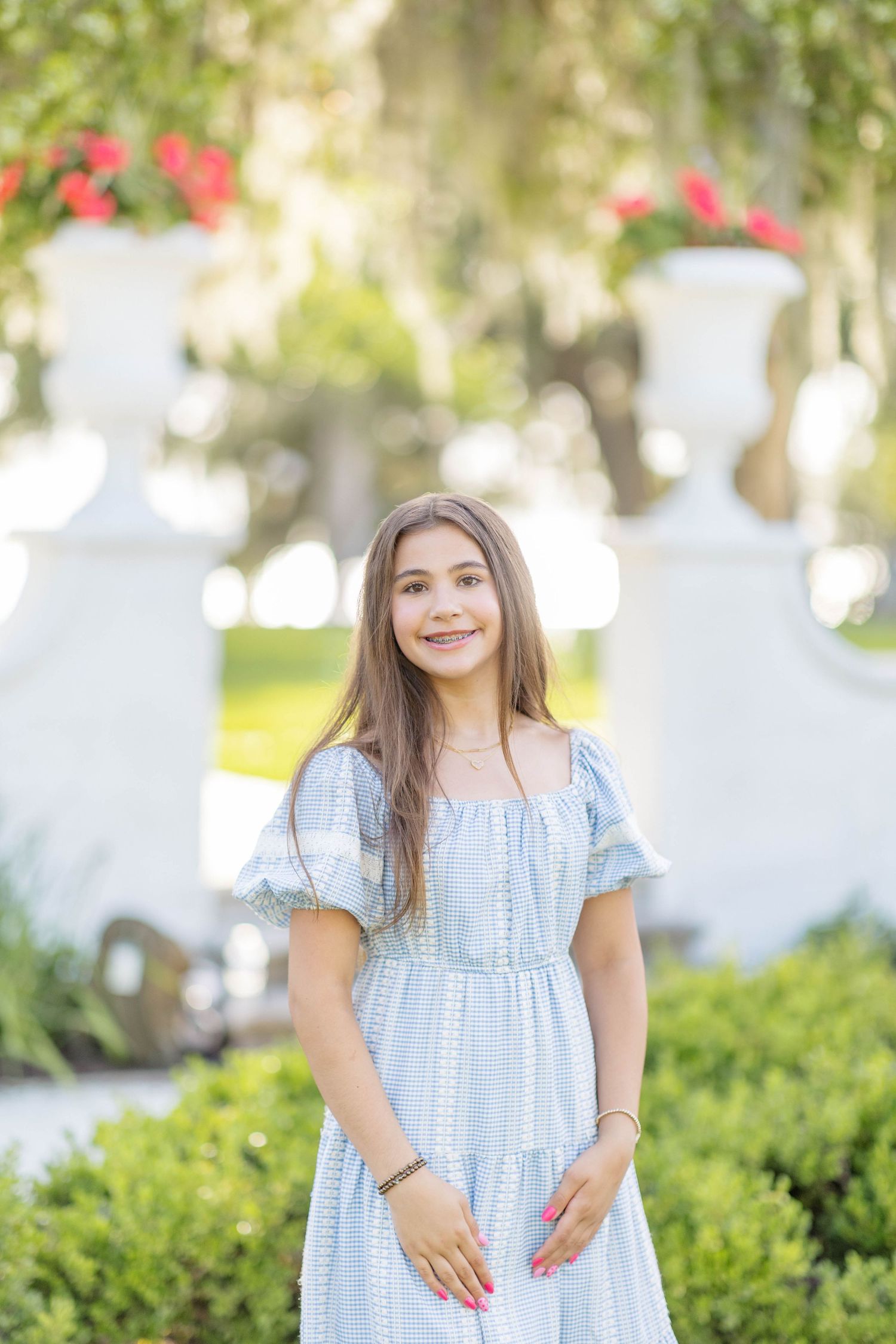 Young girl in light blue dress smiling outdoors near white decorative urns with red flowers and lush greenery.