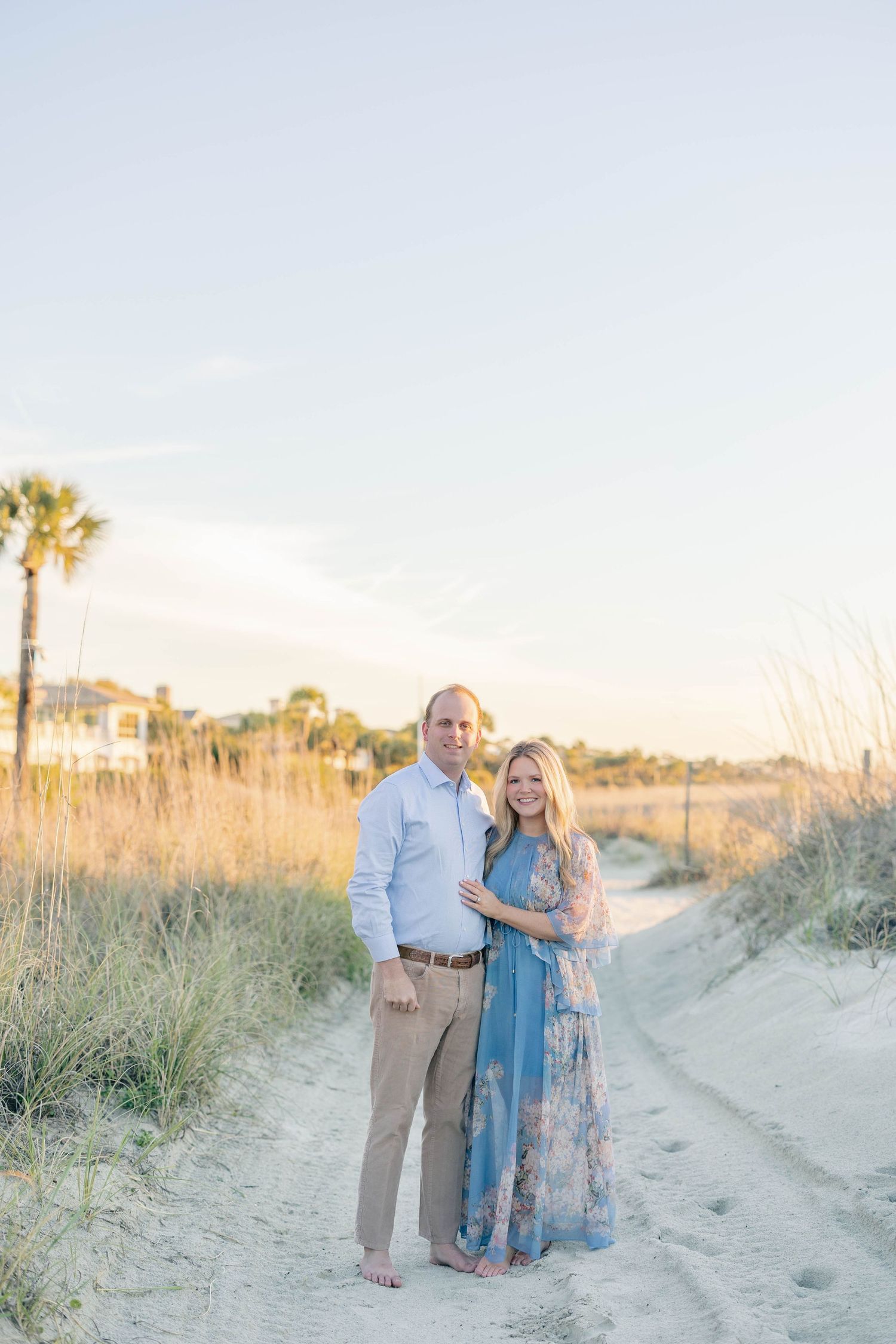 Couple posing on a sandy beach path surrounded by sea oats and palm trees during golden hour.