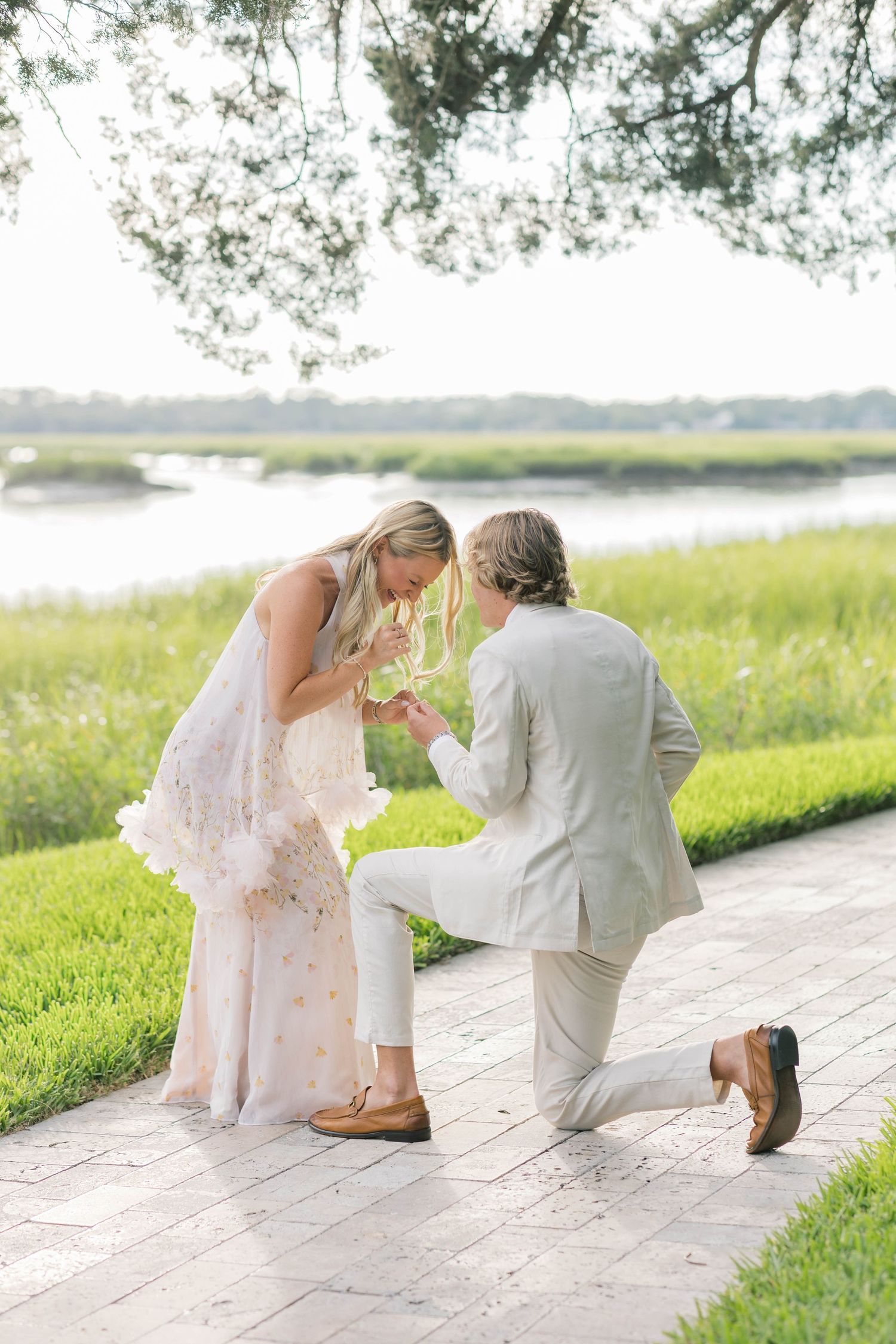 Man kneeling on one knee proposing to woman in floral dress by scenic waterway with lush green grass.