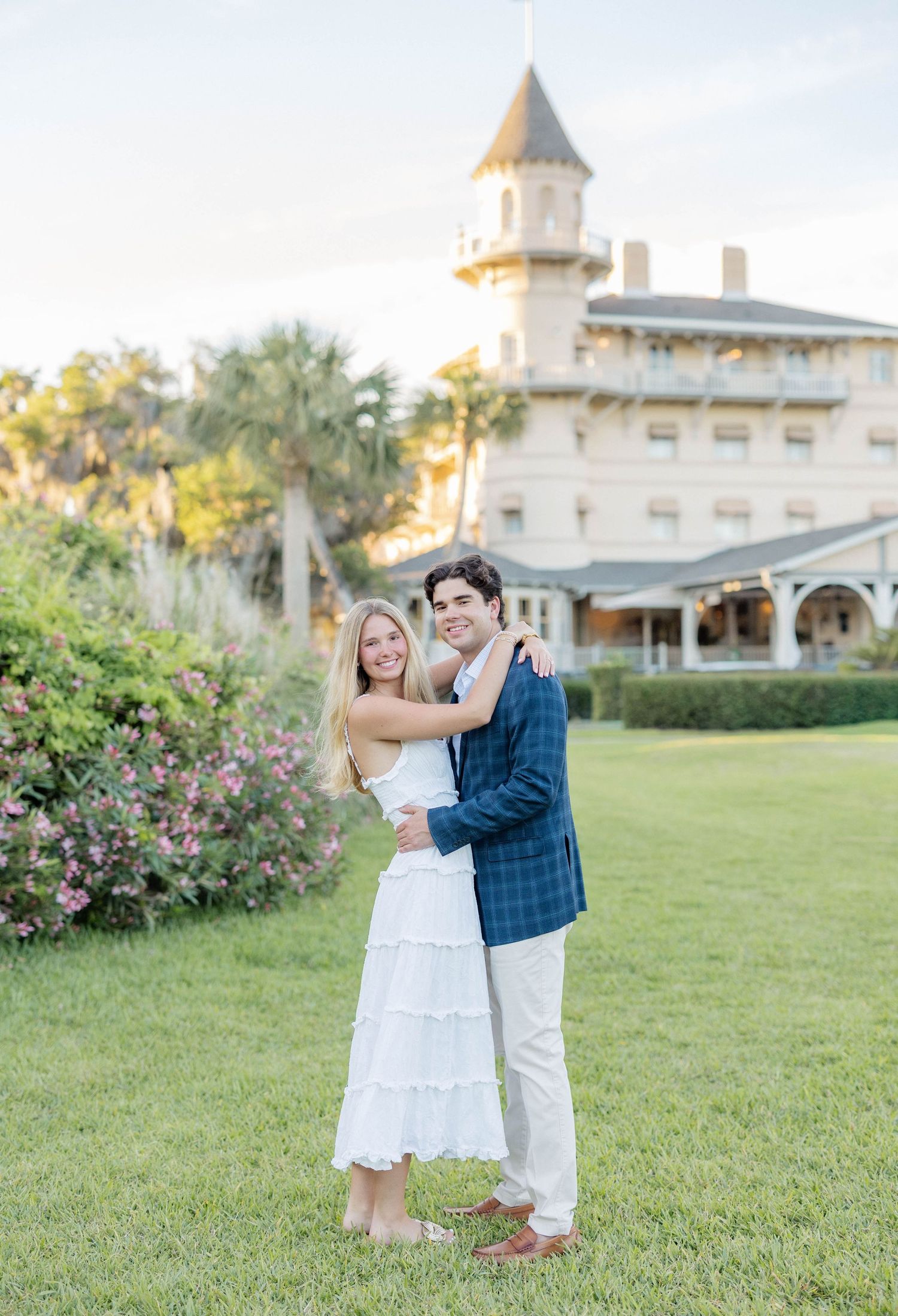 Couple embracing on green lawn before historic Victorian building with tower at golden hour.