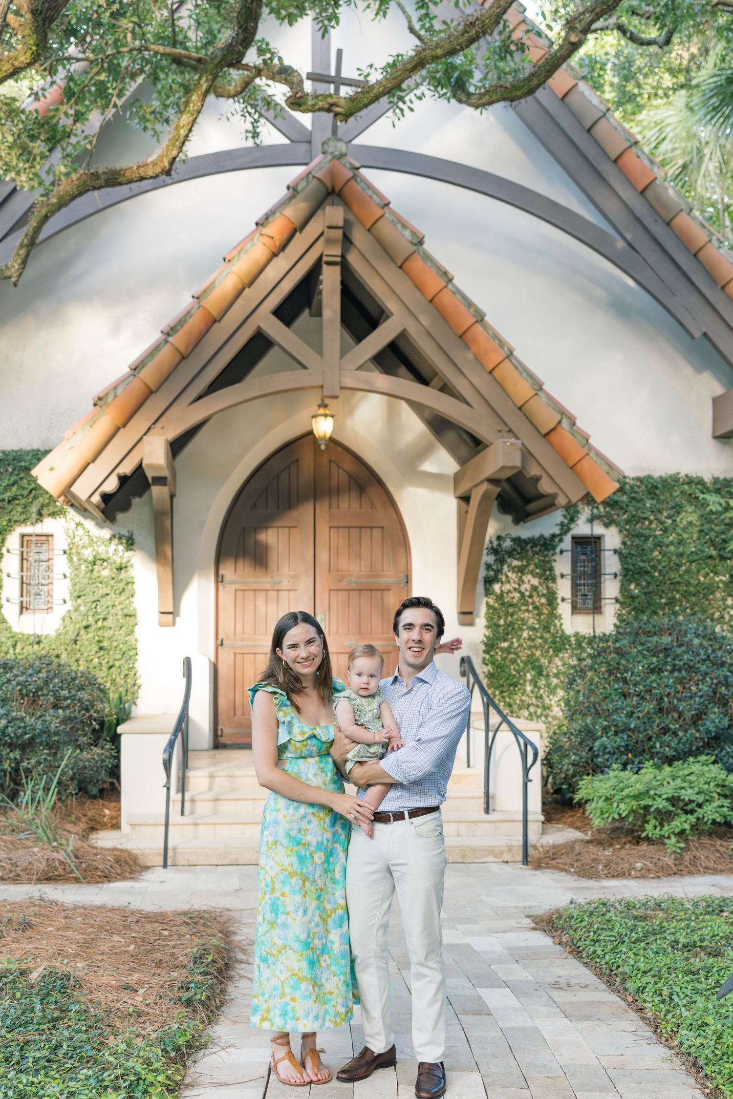 A young family poses in front of a charming chapel entrance with arched wooden beams and ivy-covered walls.
