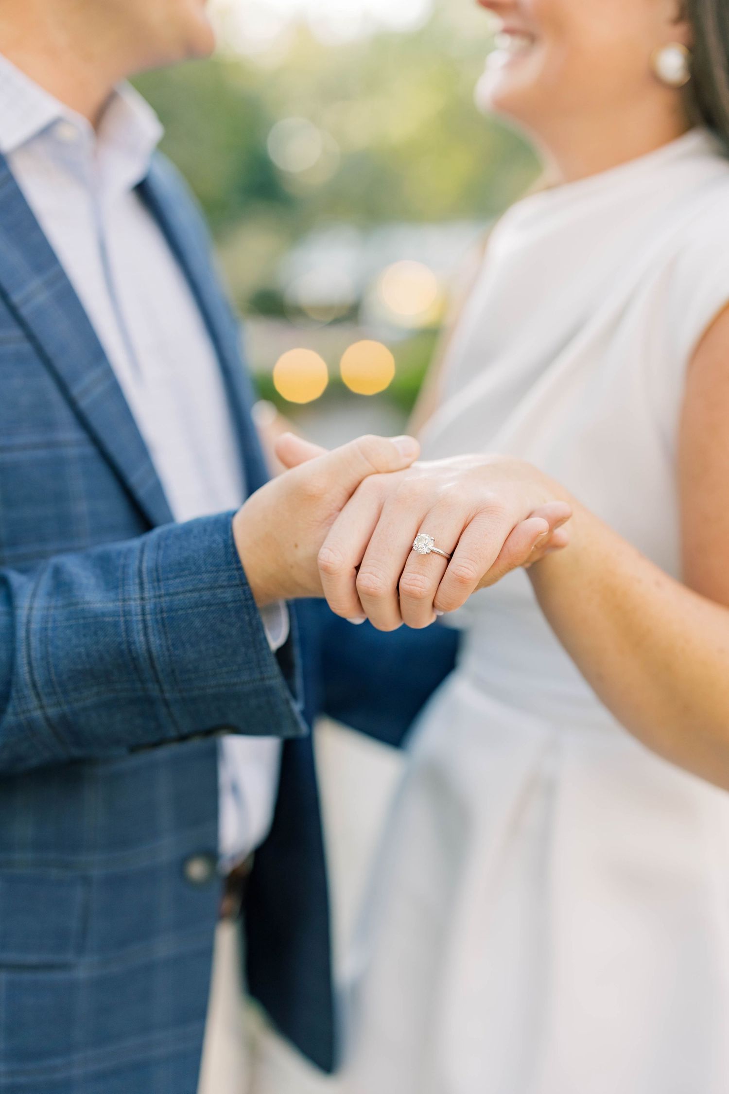 Close-up of couple holding hands showing elegant diamond engagement ring, man in blue plaid suit, woman in white dress.