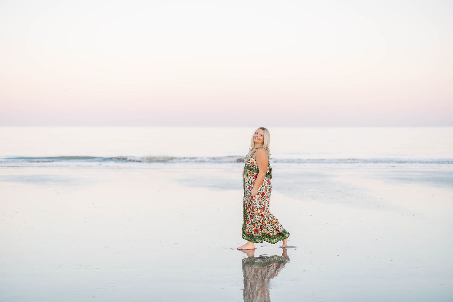 A blonde woman in a floral maxi dress stands on a reflective beach at sunset with calm ocean waves behind her.
