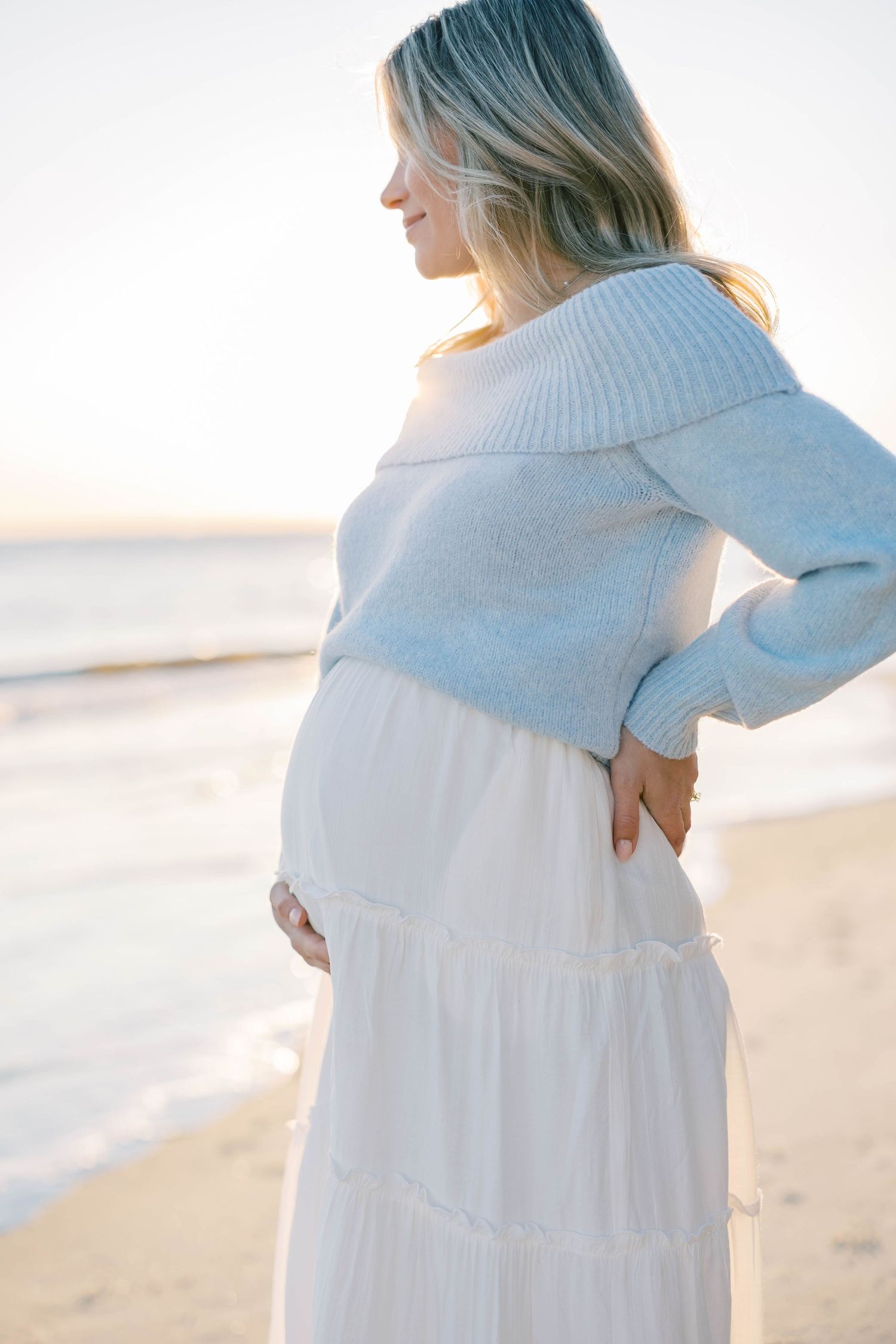 Pregnant woman in blue sweater and white skirt cradling baby bump on sandy beach at golden sunset.