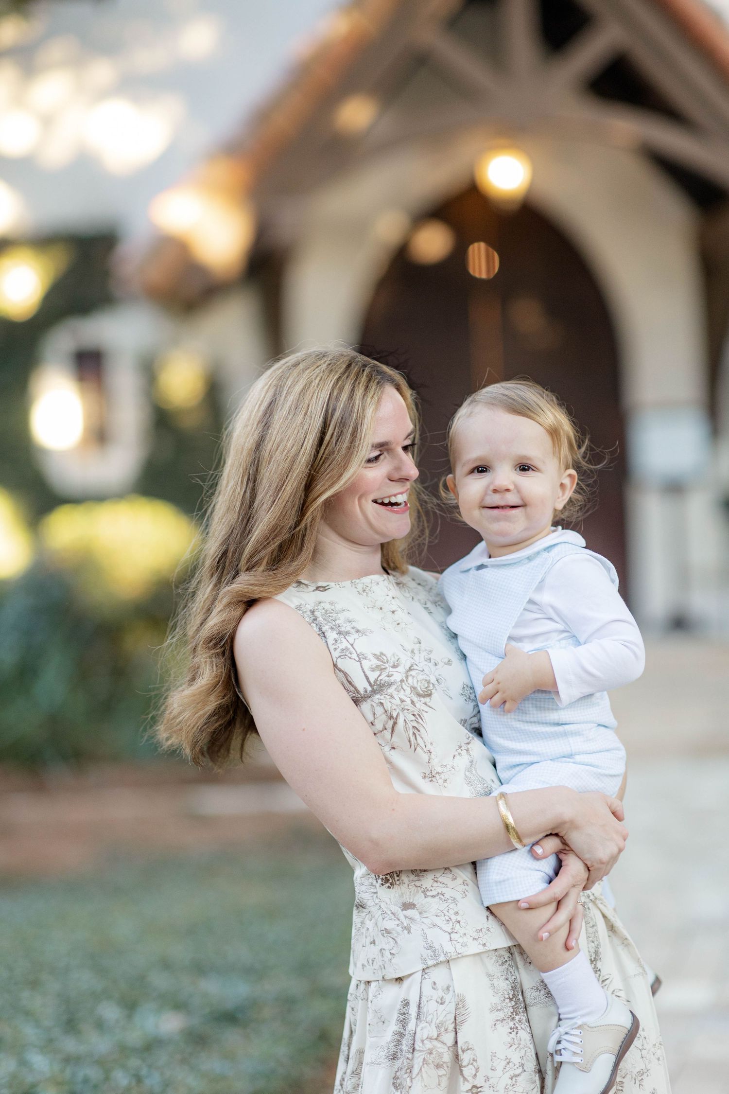 Smiling blonde woman in floral dress holding a happy toddler in white outside a charming building.