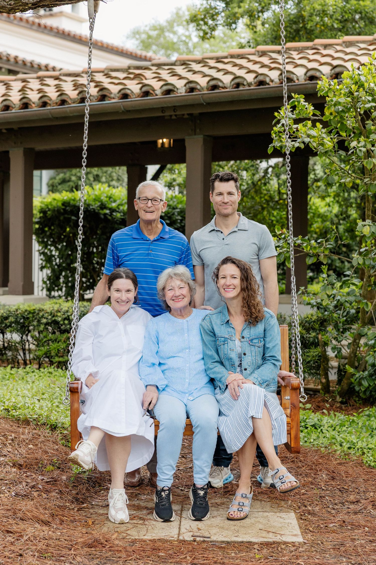 Family of five posing on a wooden porch swing surrounded by lush greenery near a terracotta-roofed building.