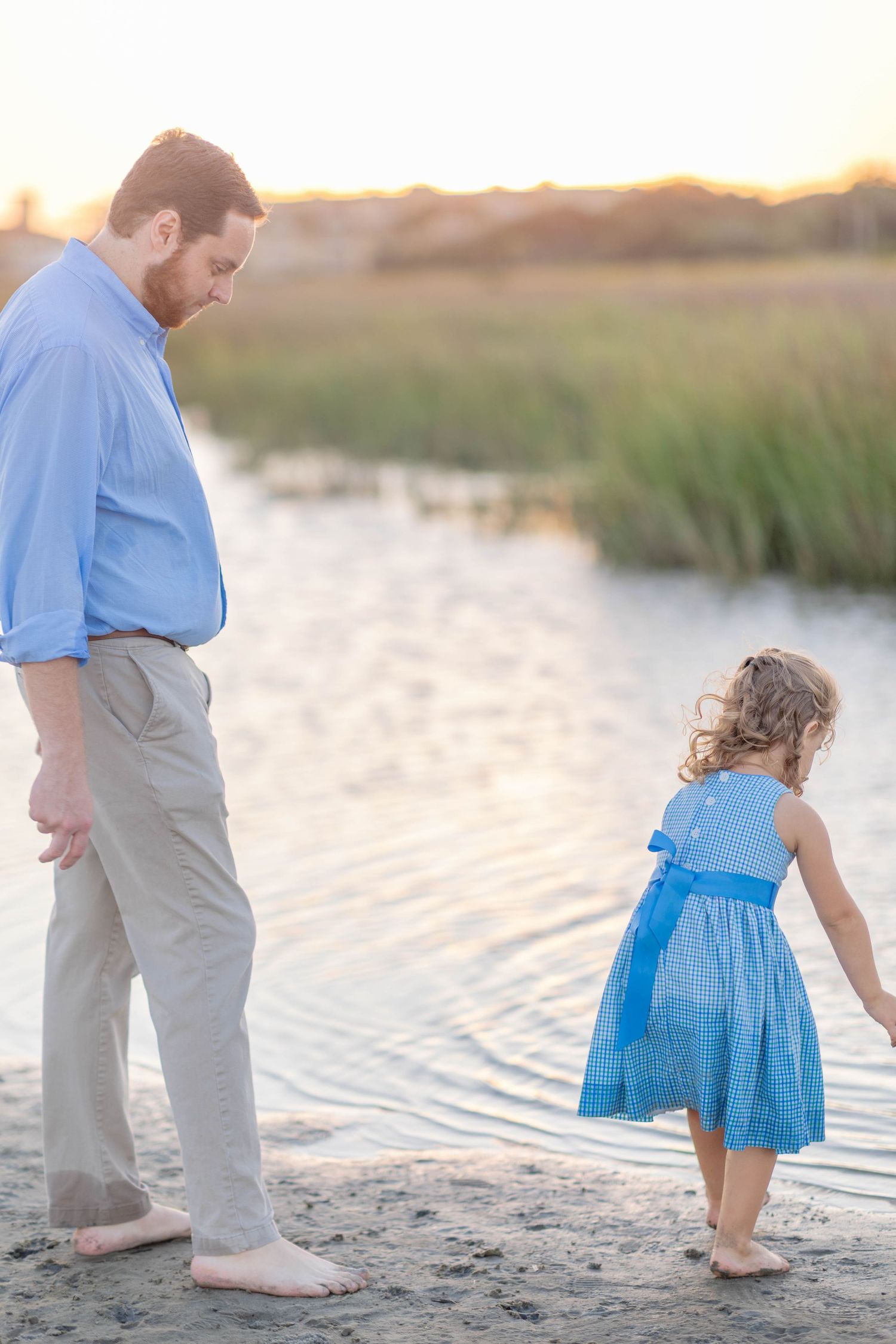 A bearded father in a blue shirt walks barefoot with his young daughter in a blue dress by a serene waterway at sunset.