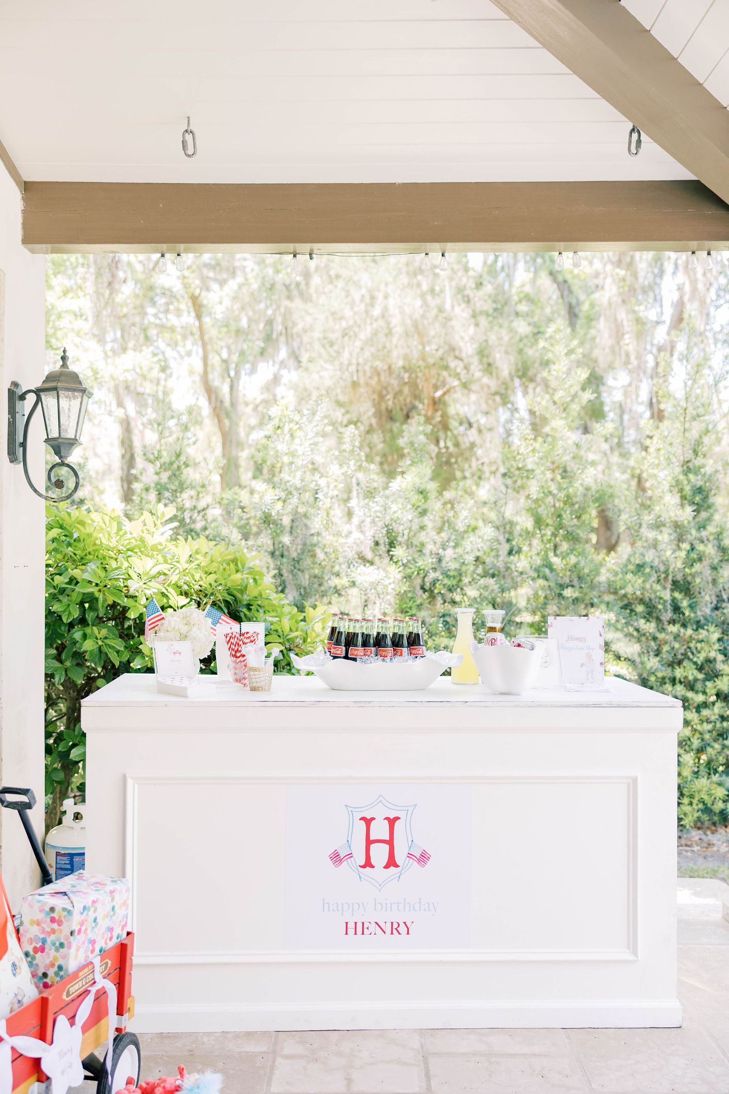 White bar setup with Henry monogram sign, mini Coca-Cola bottles, and floral arrangements at an outdoor party.