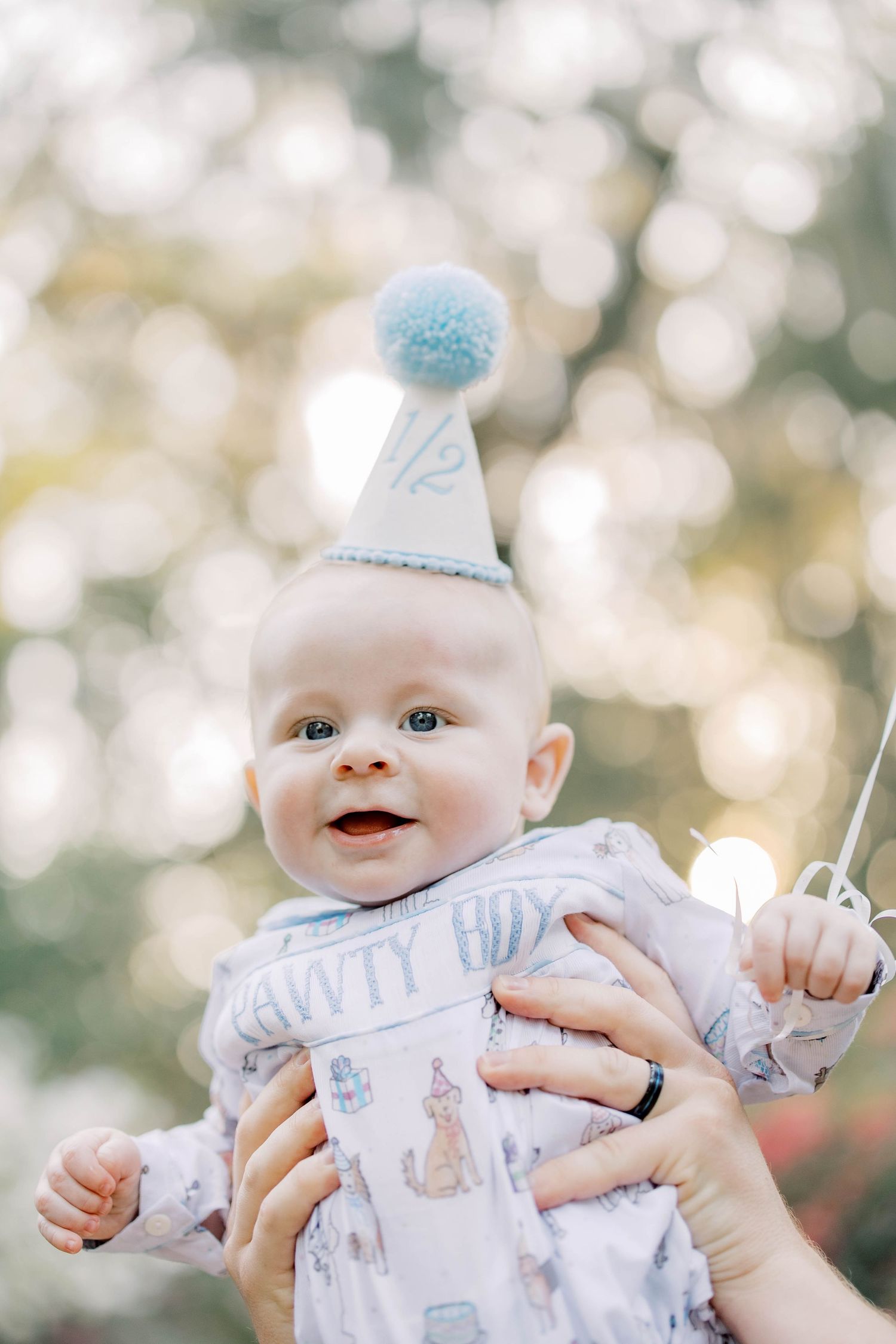 Smiling baby wearing a blue pom-pom half birthday hat, held up outdoors with bokeh background.