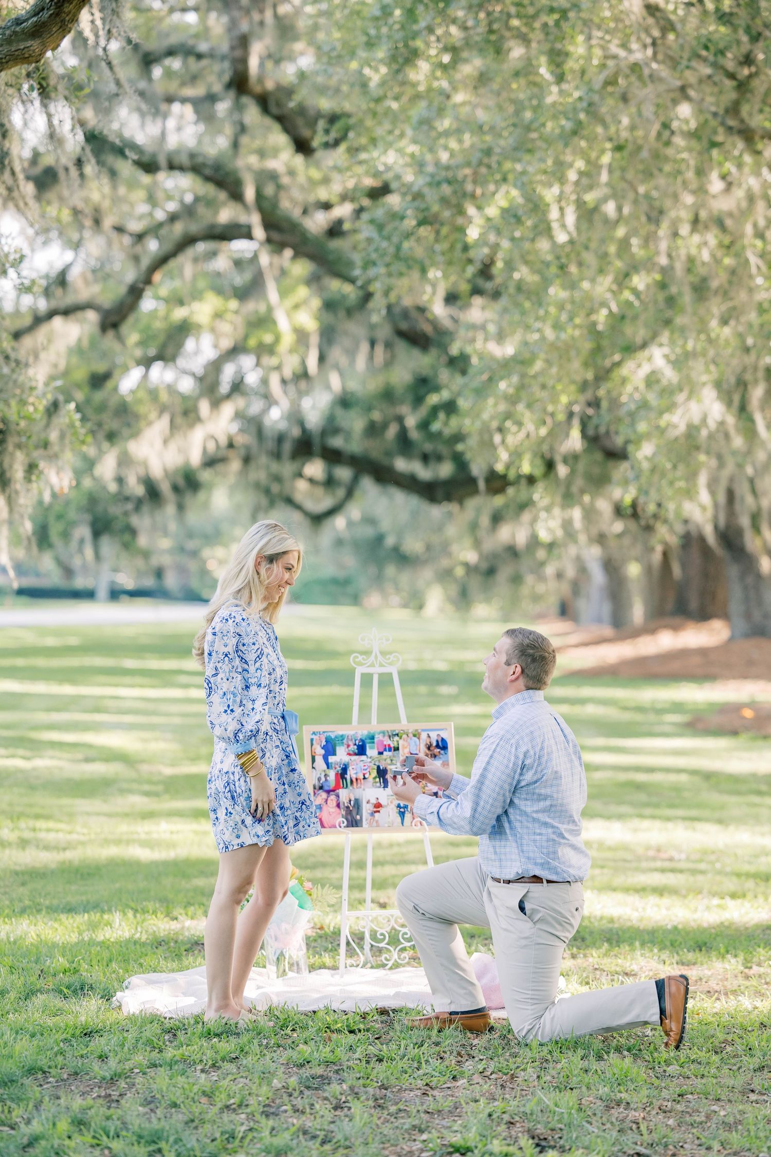 Man kneeling on one knee proposing to woman in blue floral dress outdoors under oak trees with photo display on easel.