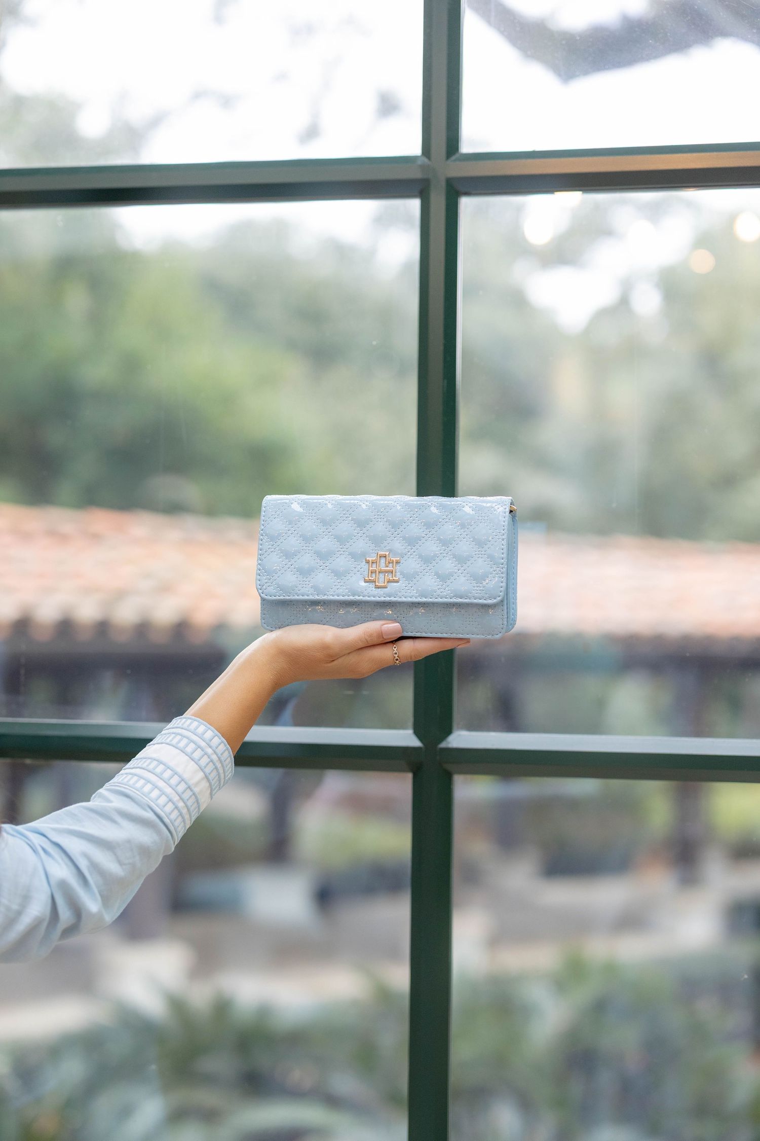 Hand holding a light blue quilted Tory Burch wallet with gold logo clasp against a green-framed window.