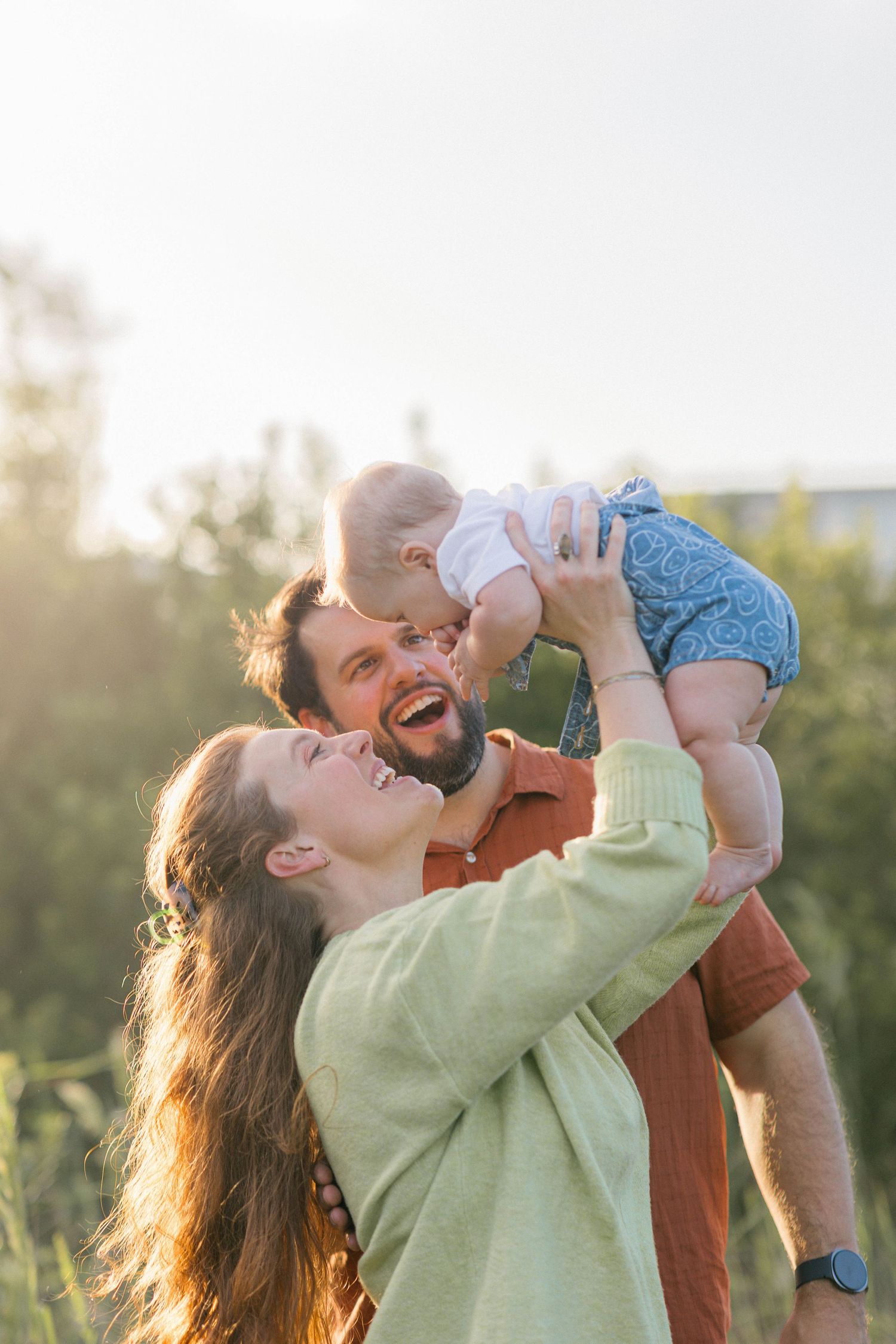 Happy young family with baby outdoors in sunlight, parents laughing as father lifts infant up in green nature setting.