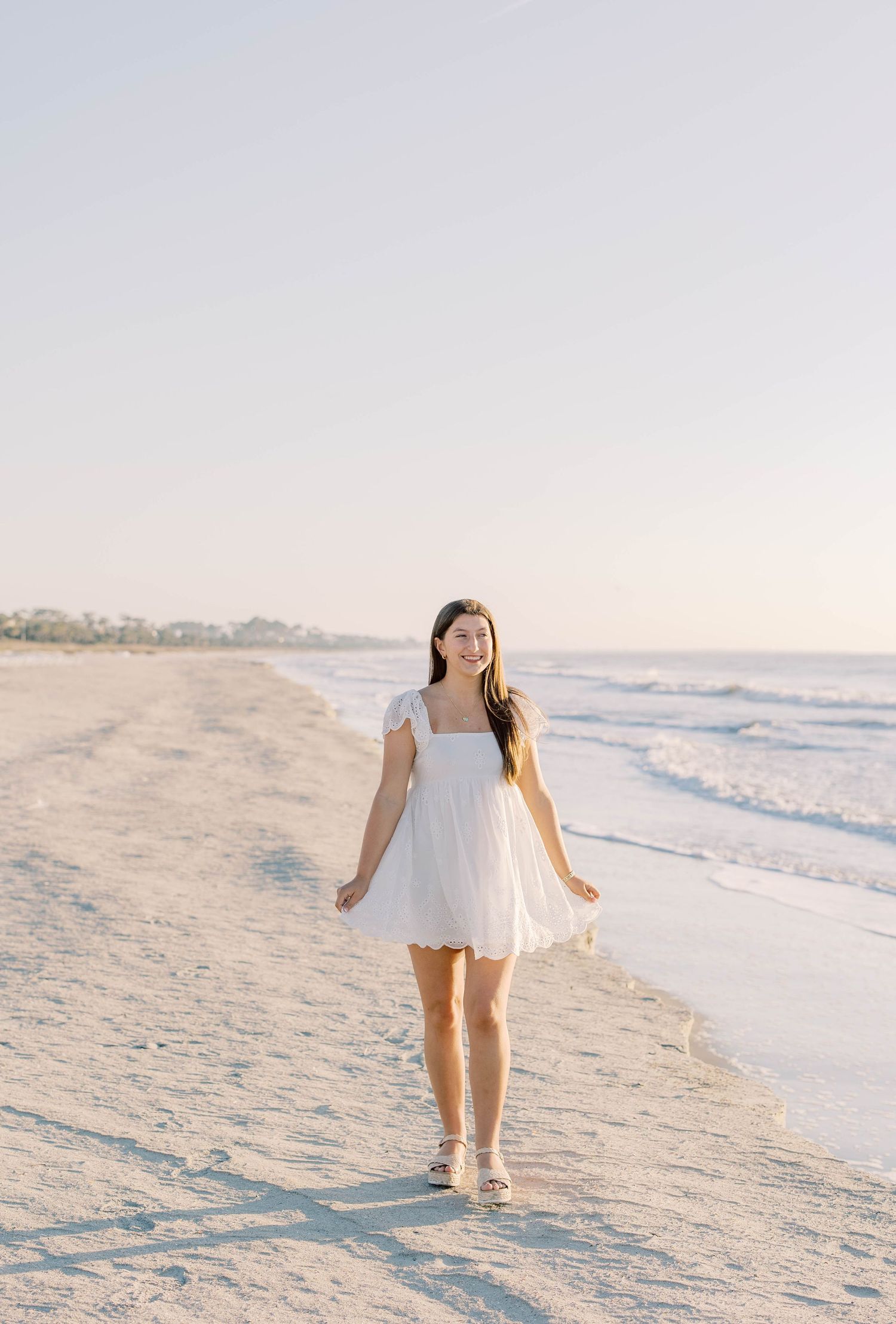 A young woman in a white mini dress walks along a sunny sandy beach with ocean waves in the background.