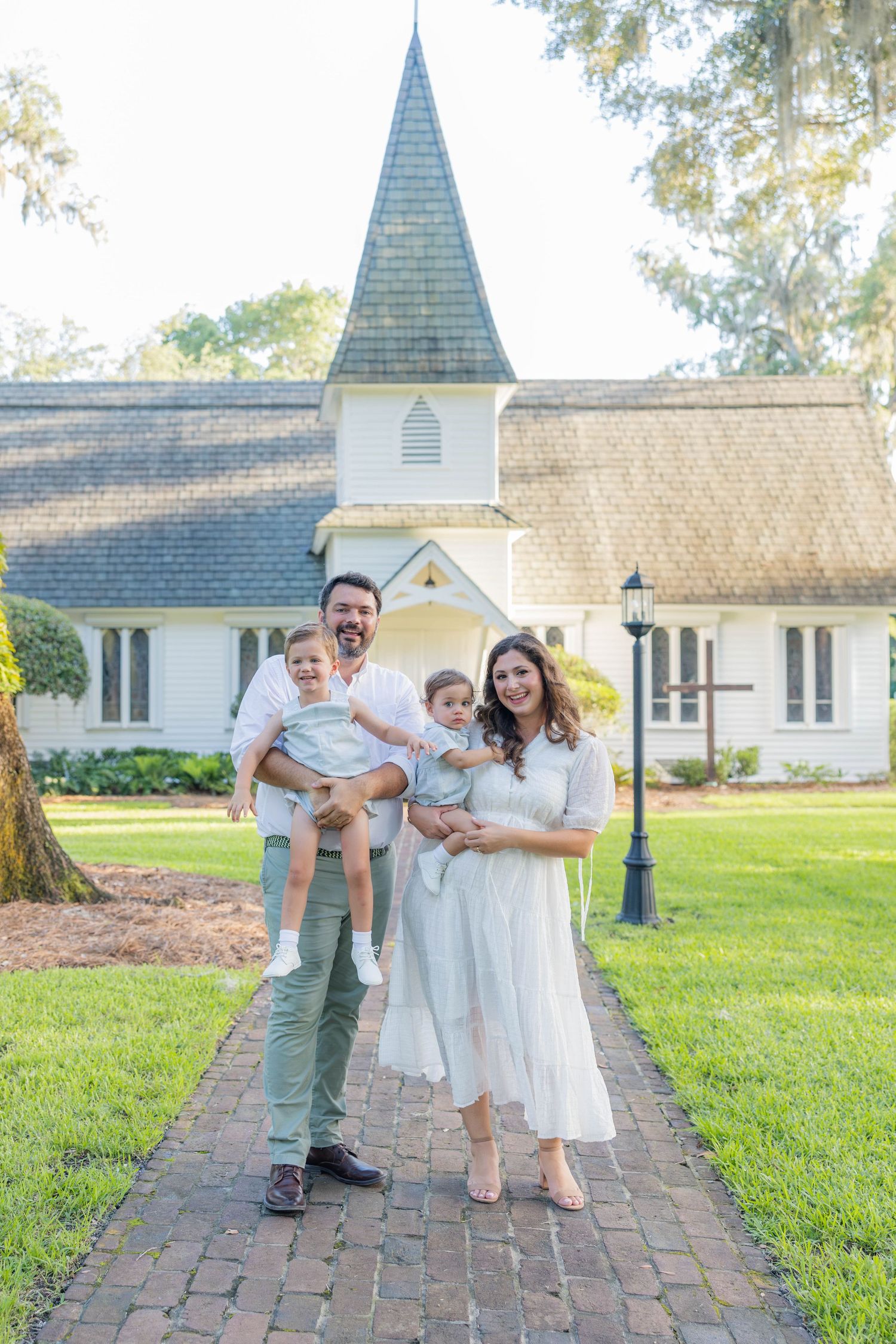 Family of four posing on brick path before white chapel, parents holding two young children, dressed in white and pastels.