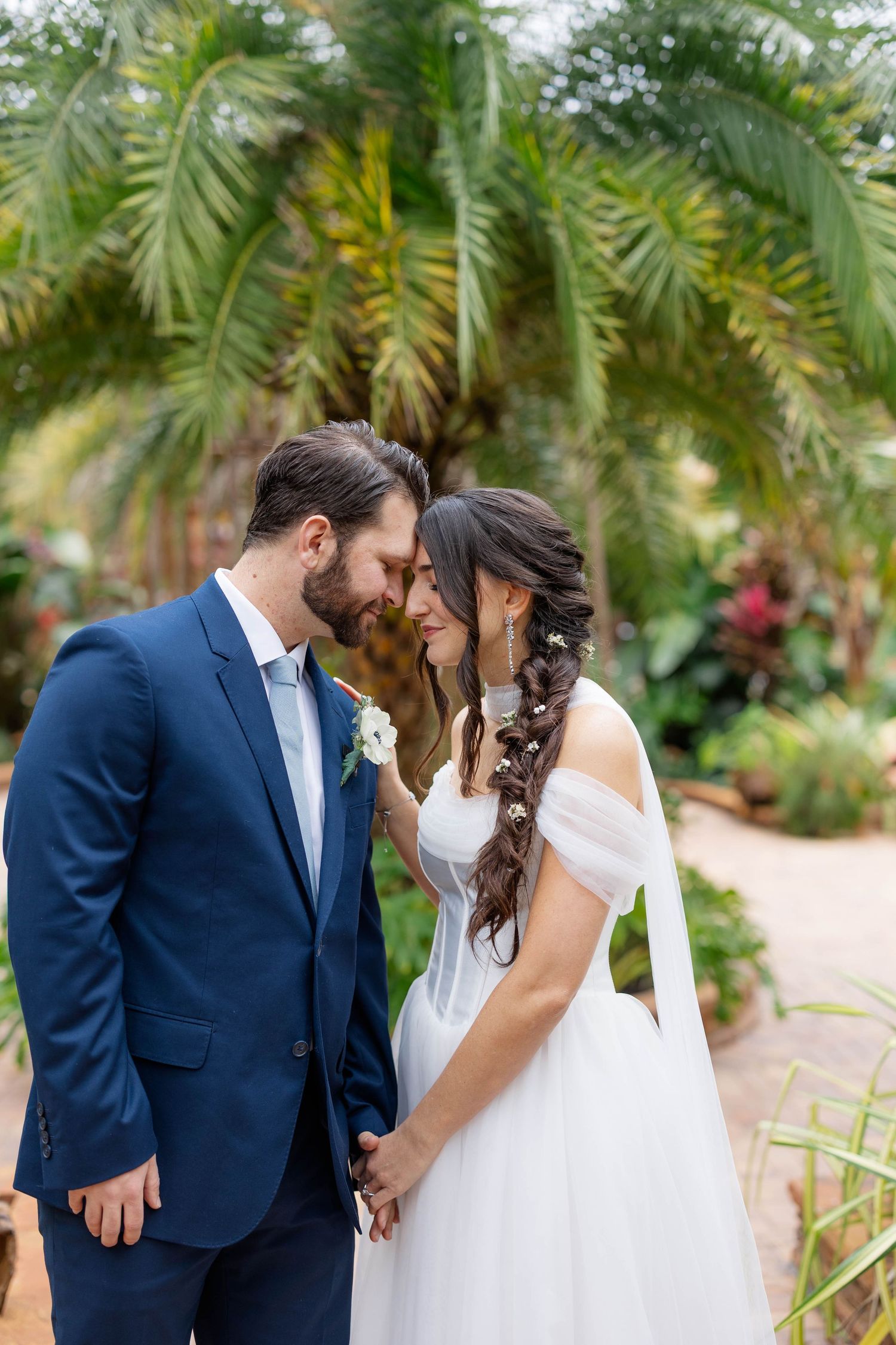 A romantic couple in wedding attire share intimate moments among tropical palm trees in an outdoor garden setting.
