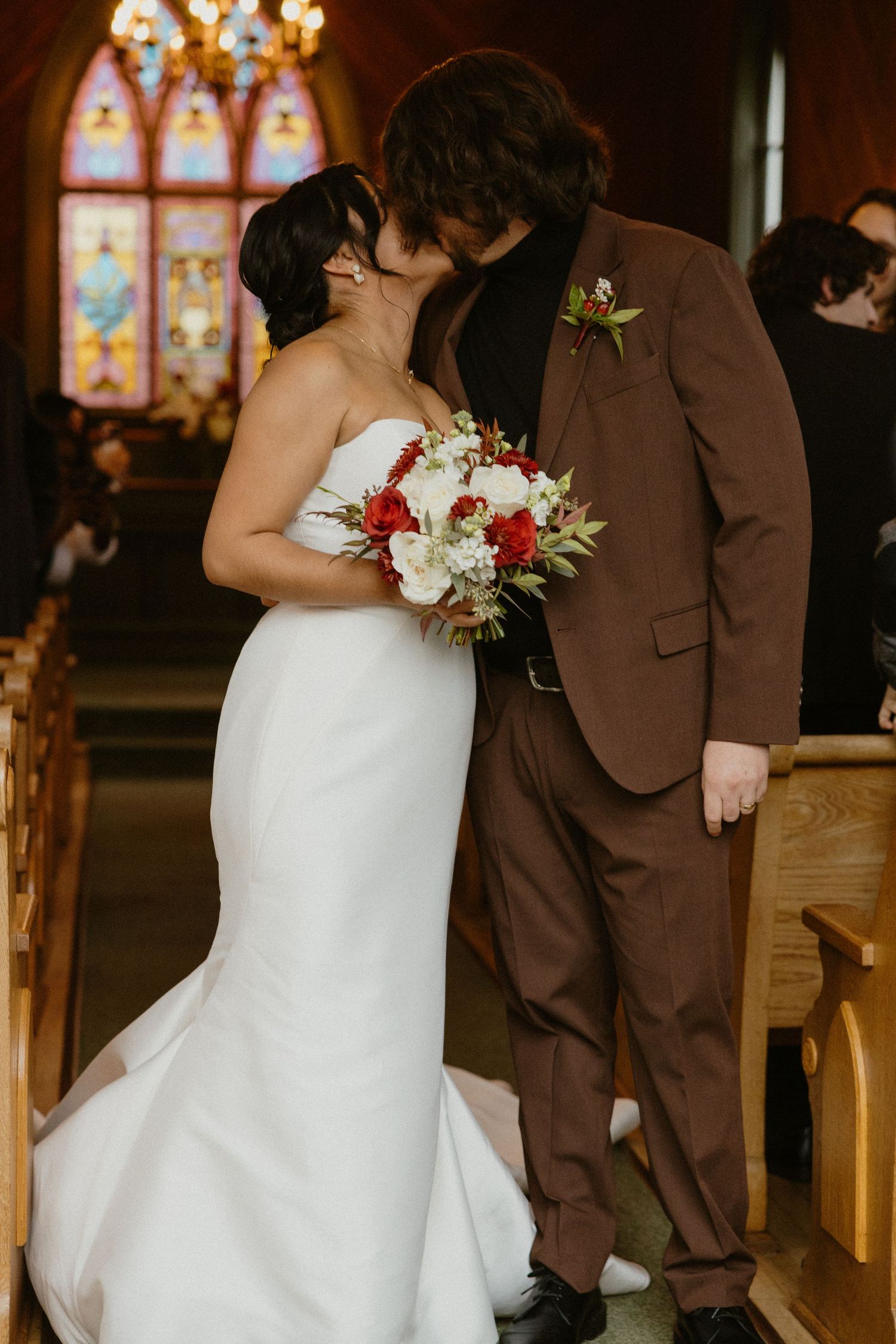 Bride and groom share first kiss in church aisle with stained glass windows in background.
