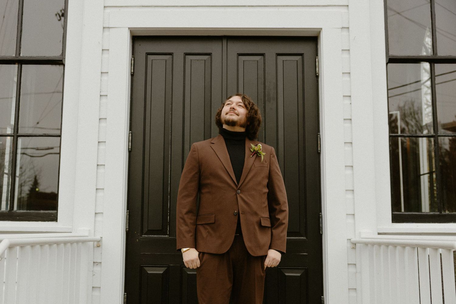 Groom in brown suit with boutonniere standing confidently before a black double door entrance.
