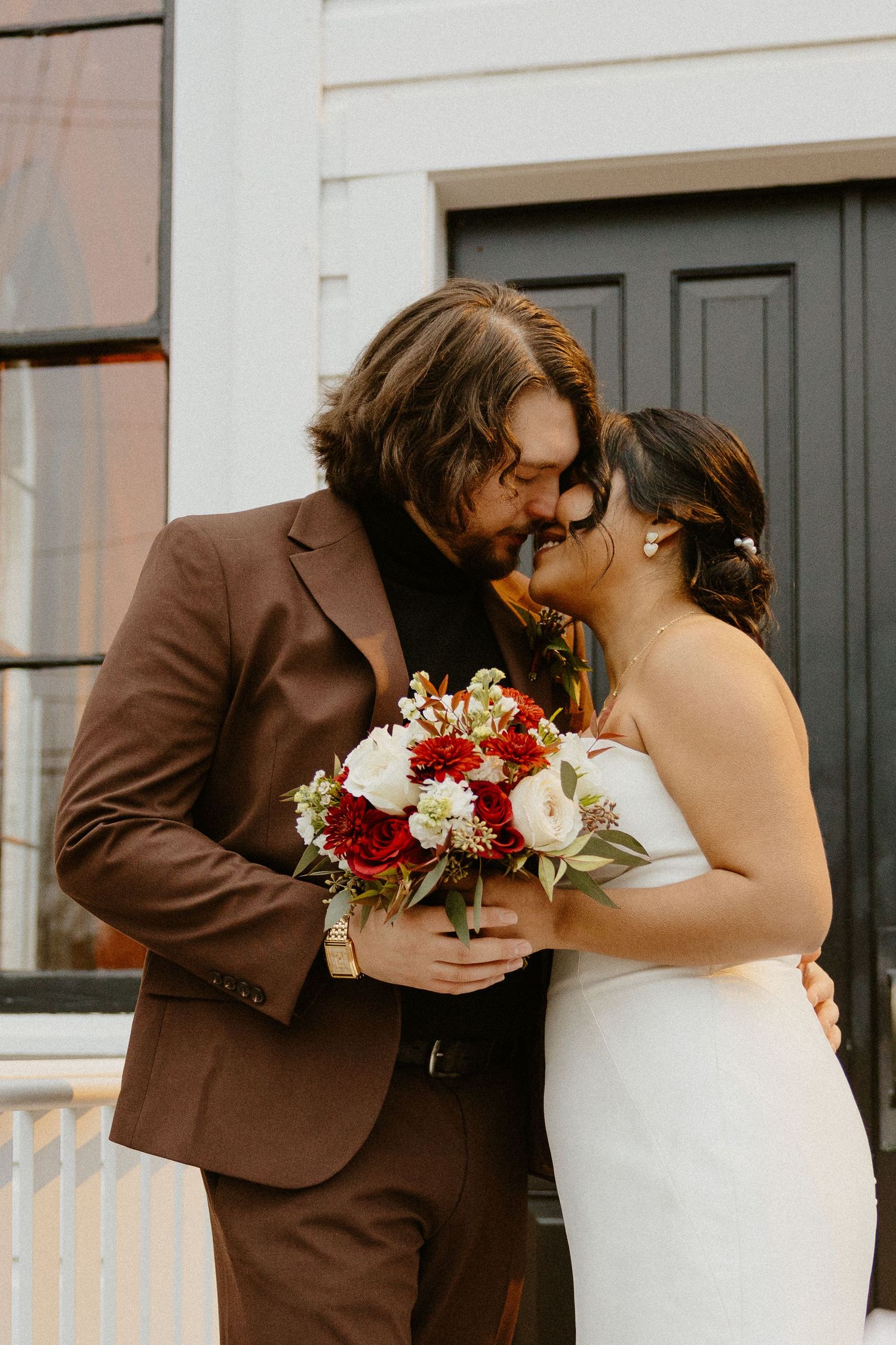 Bride and groom kissing, holding red and white floral bouquet, groom in brown suit.