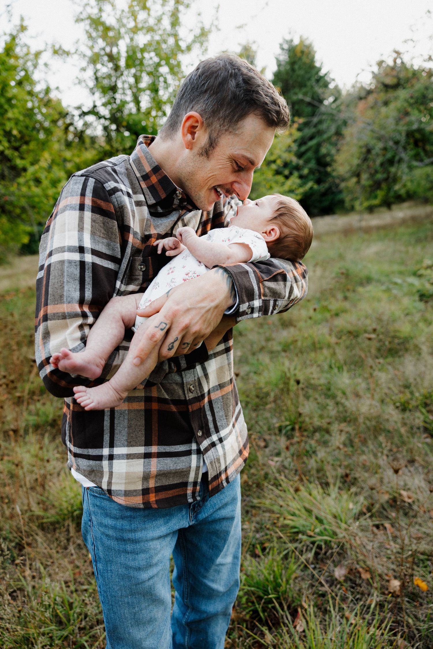 A smiling father in a plaid shirt joyfully nuzzles his newborn baby outdoors in a green field.