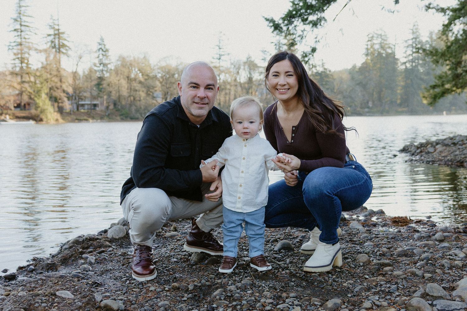 Family of three crouching together on rocky lakeshore, smiling, with evergreen trees in the background.