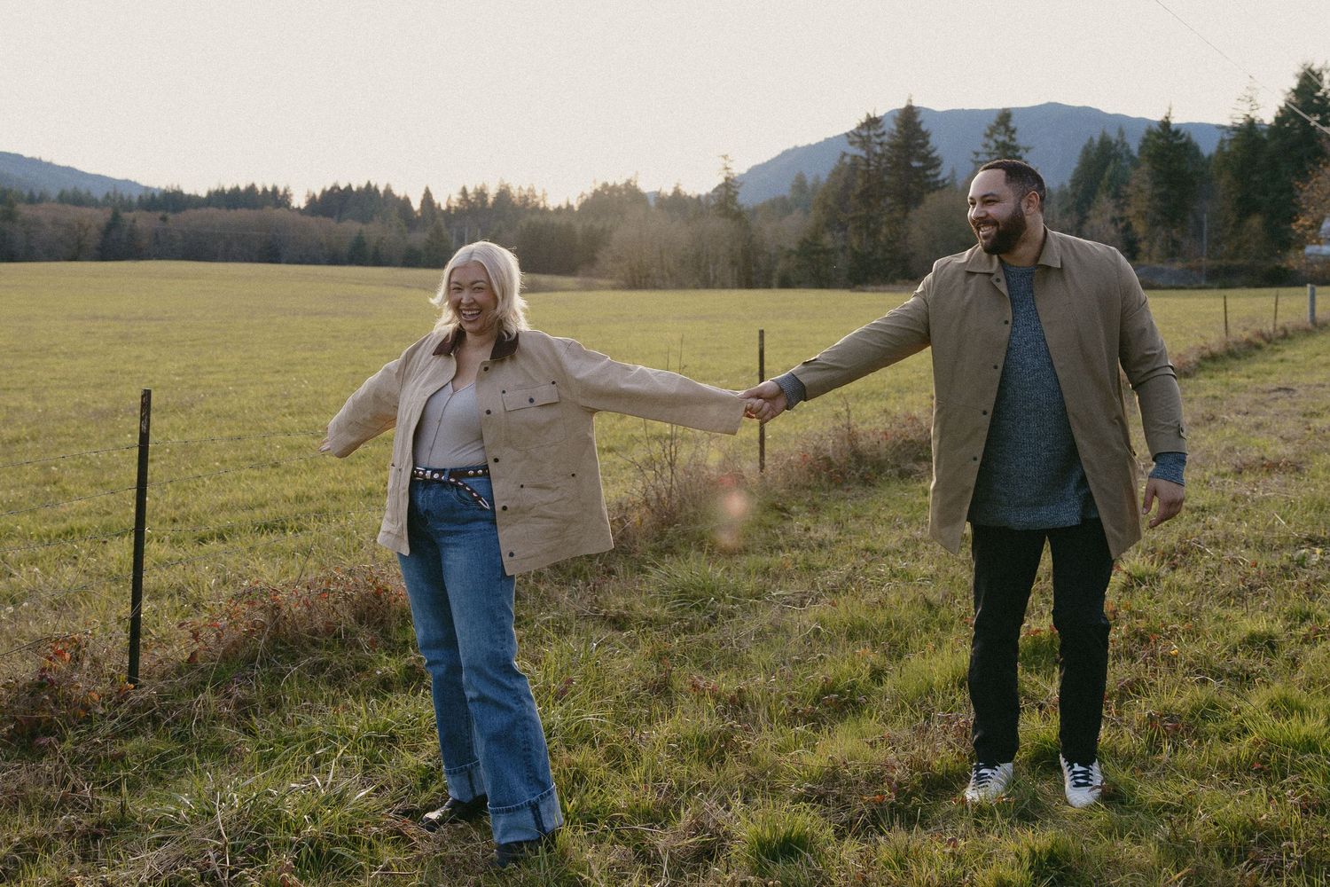 Couple holding hands walking through green field at golden hour sunset.