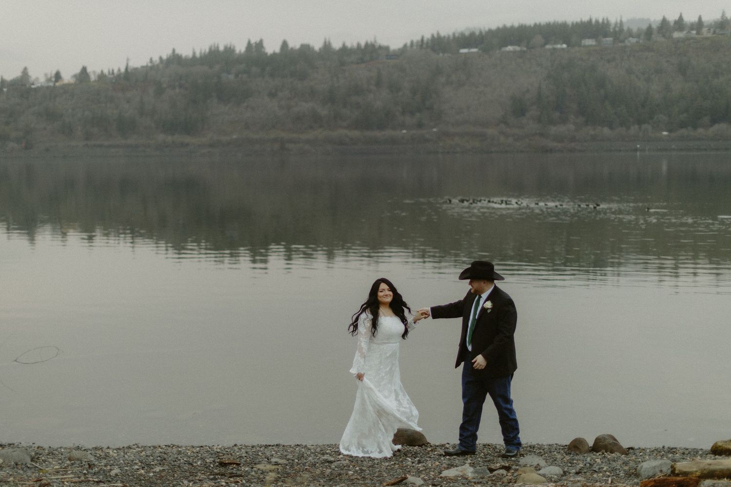 Bride in white lace gown and groom in black cowboy hat walk hand in hand along a rocky lakeside shore.