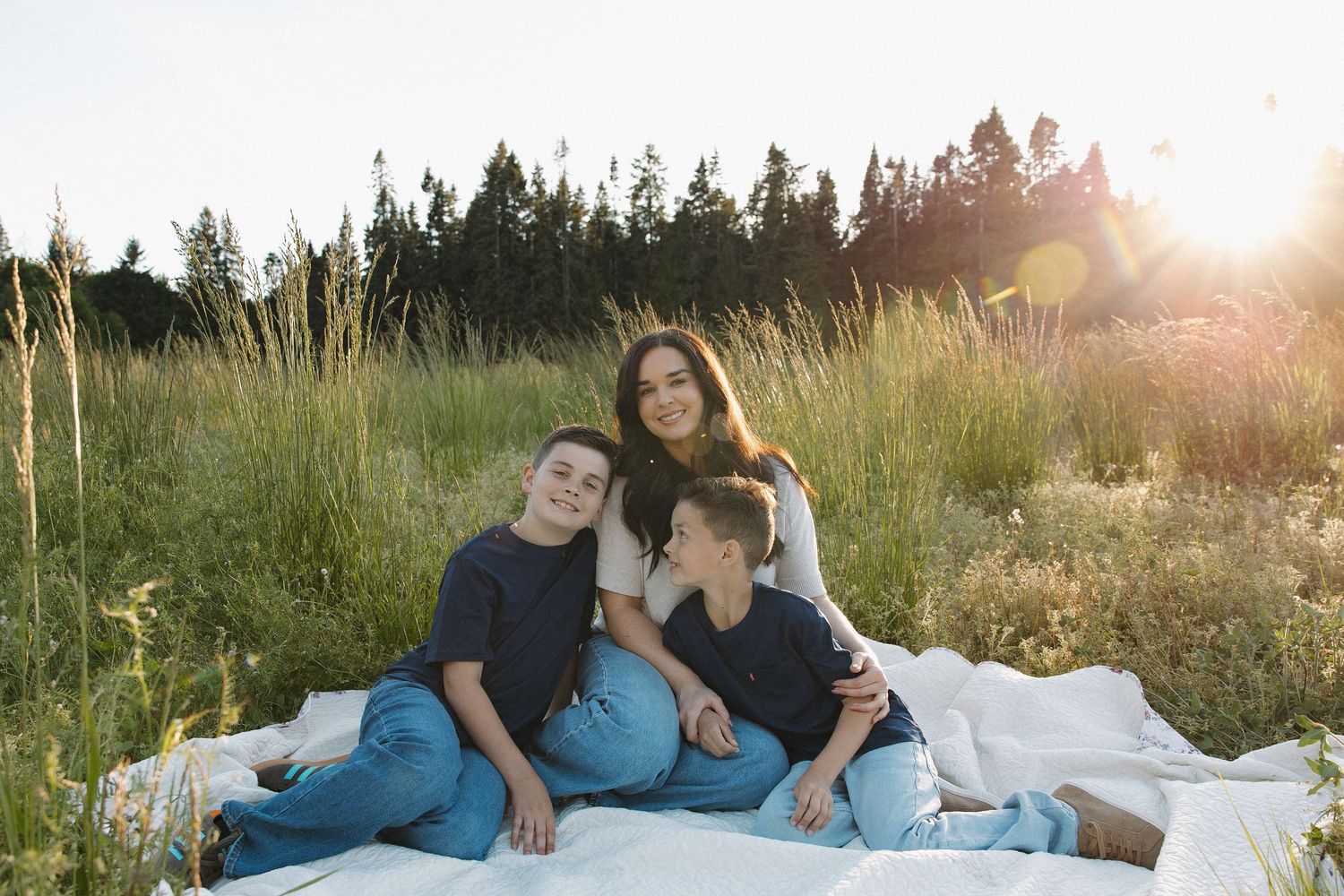 A mother sits on a white blanket in a sunny meadow, smiling with her two young sons surrounded by tall grass.