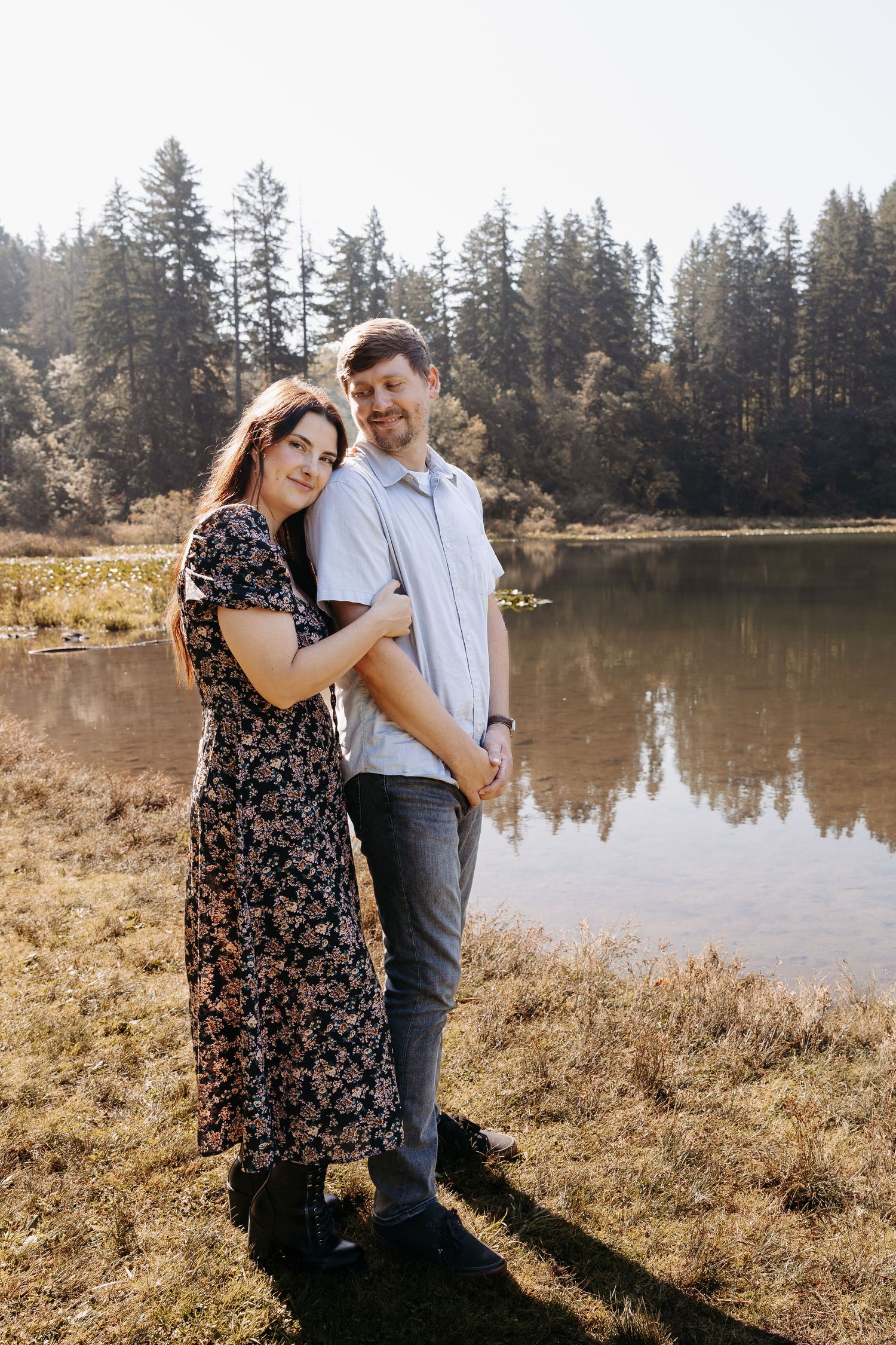 Couple embracing by a reflective lake surrounded by pine trees.