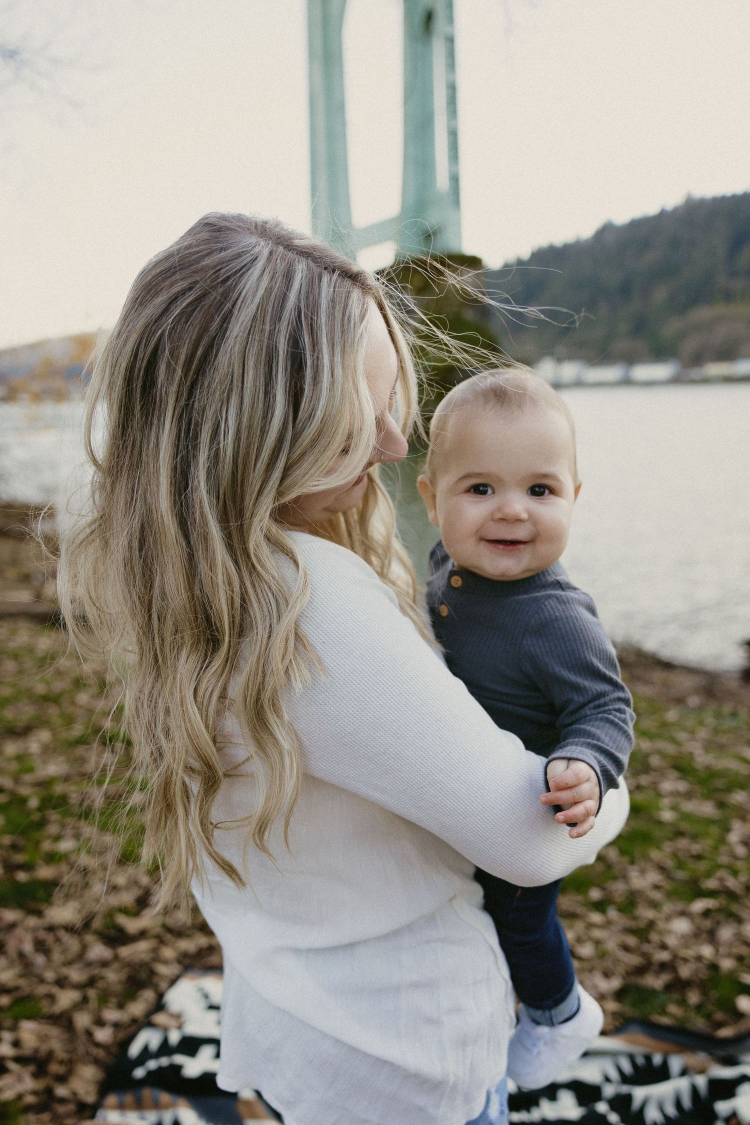 Blonde mother holding smiling baby boy near river with bridge in background.