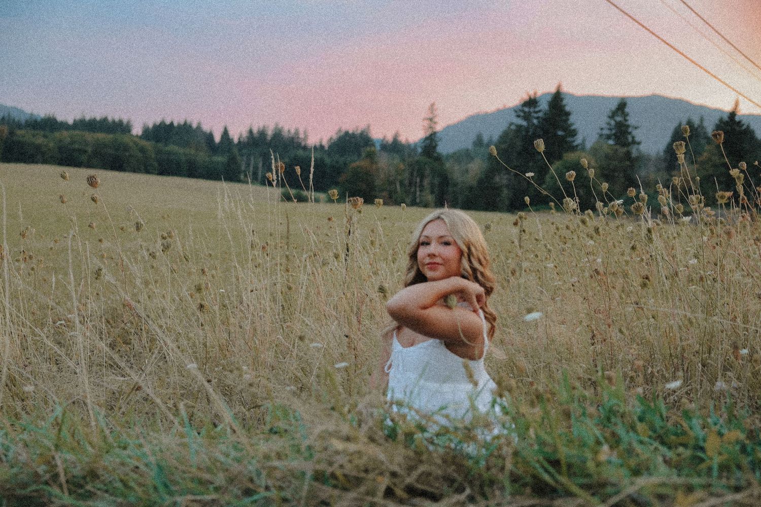 Young woman sitting in golden field hugging herself at sunset with mountains behind.