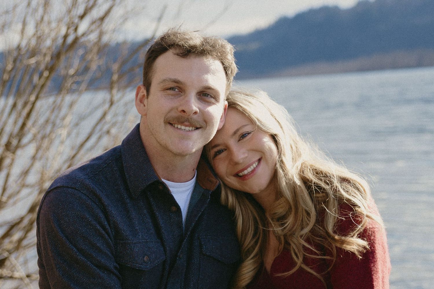 Couple smiling warmly together near a lake with bare branches, wearing casual fall-colored outfits.
