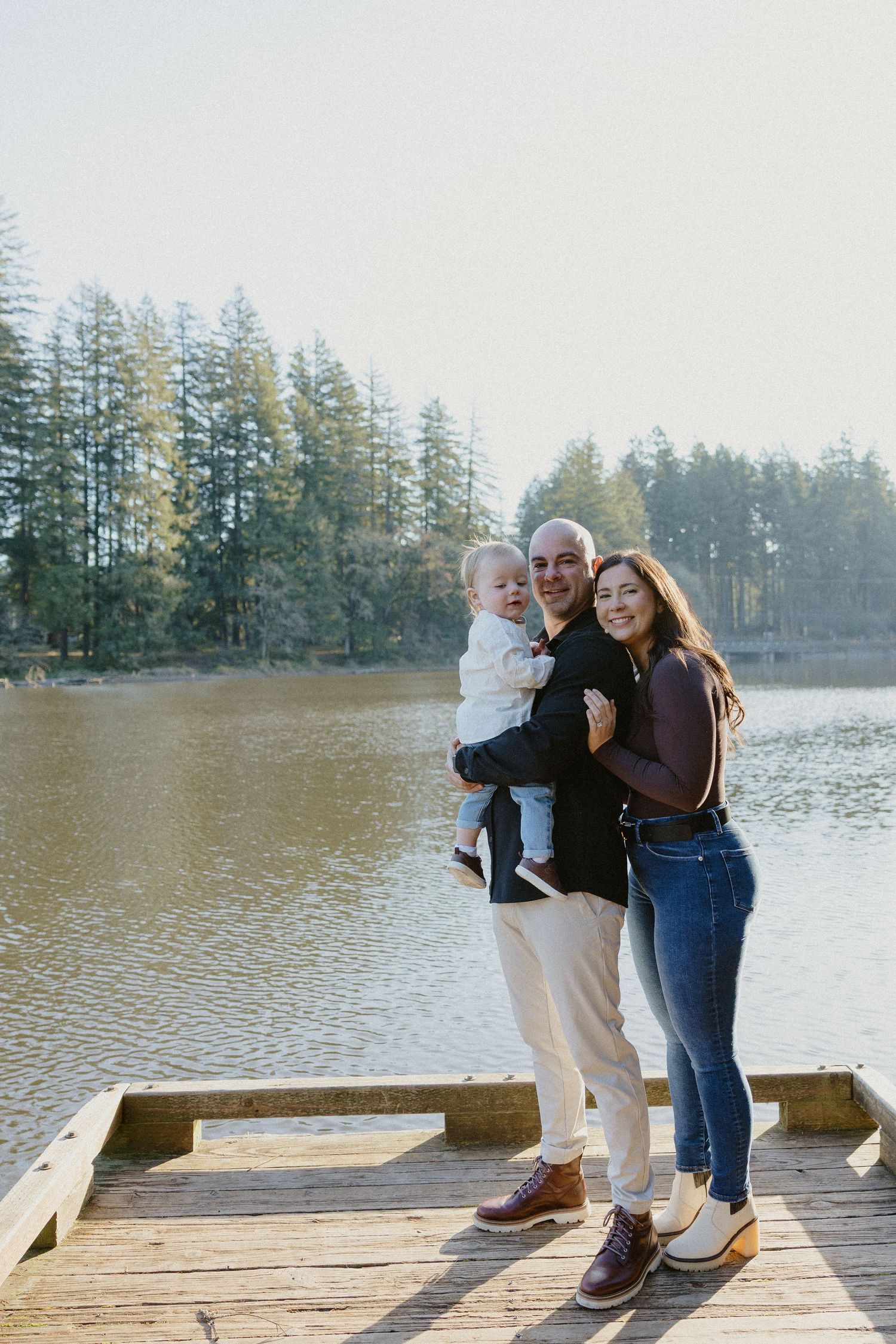 Smiling family of three posing on a wooden dock with a serene lake and forest behind them.