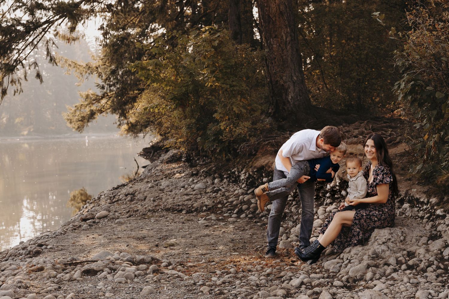 Family of four relaxing on rocky riverbank by calm water, parents engaging with two children.