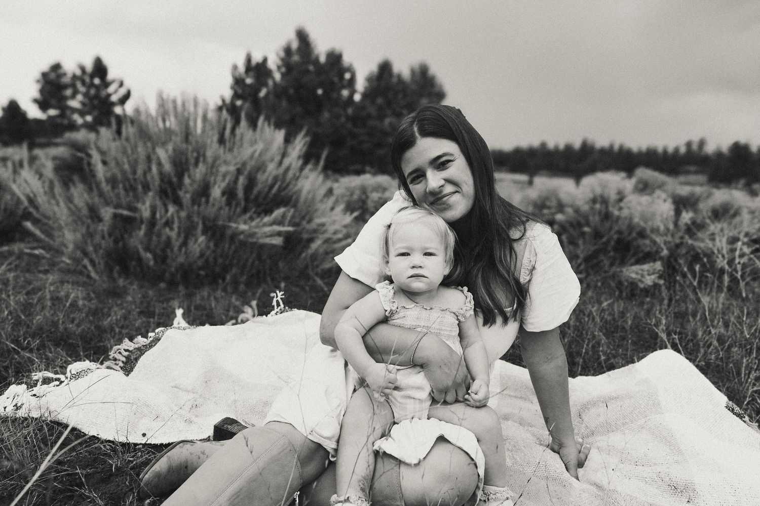 Black and white portrait of smiling mother sitting with baby girl on blanket in outdoor field.