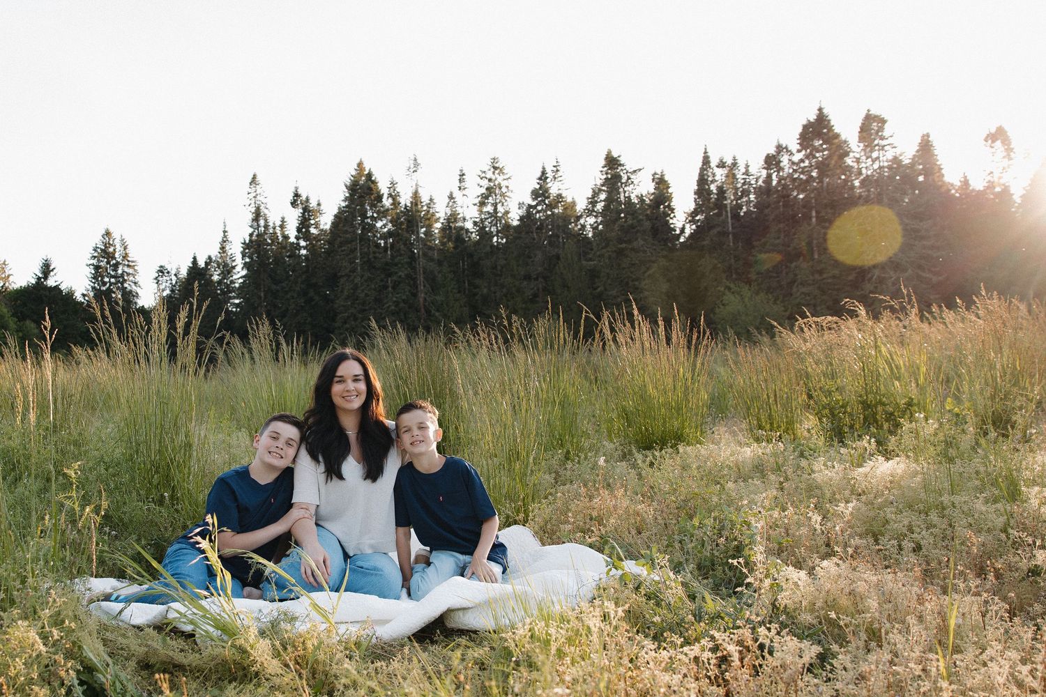 A mother sits with her two children on a blanket in a sunlit meadow with evergreen trees in the background.