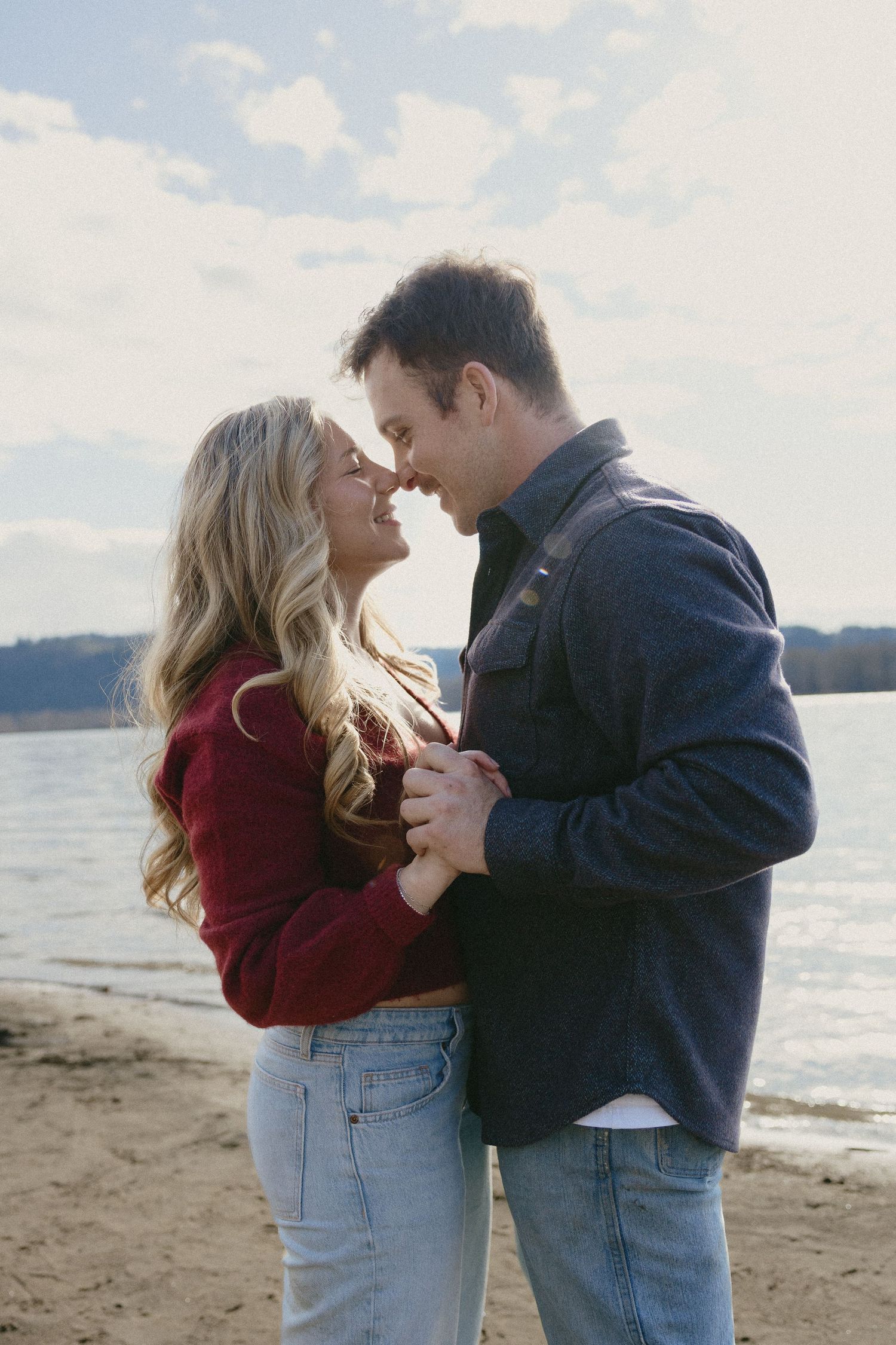 Couple smiling and holding each other close on a sunny beach by a lake in fall outfits.